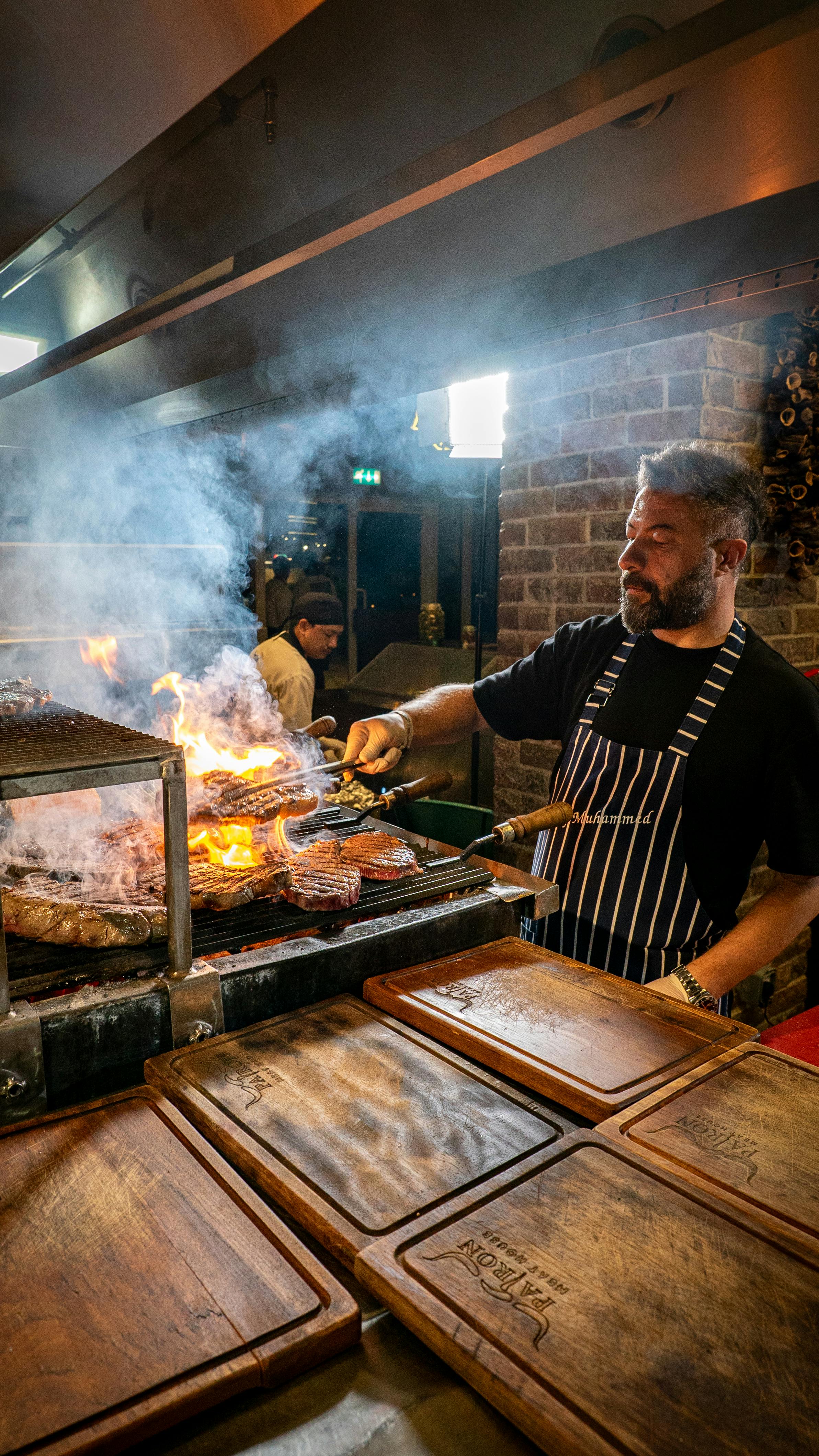 A grill master cooking thick steaks over open flames in a rustic wood-fired kitchen