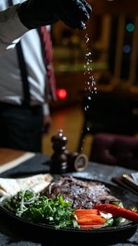 Chef sprinkles seasoning on gourmet steak dish with fresh vegetables in a dimly lit, elegant restaurant setting.