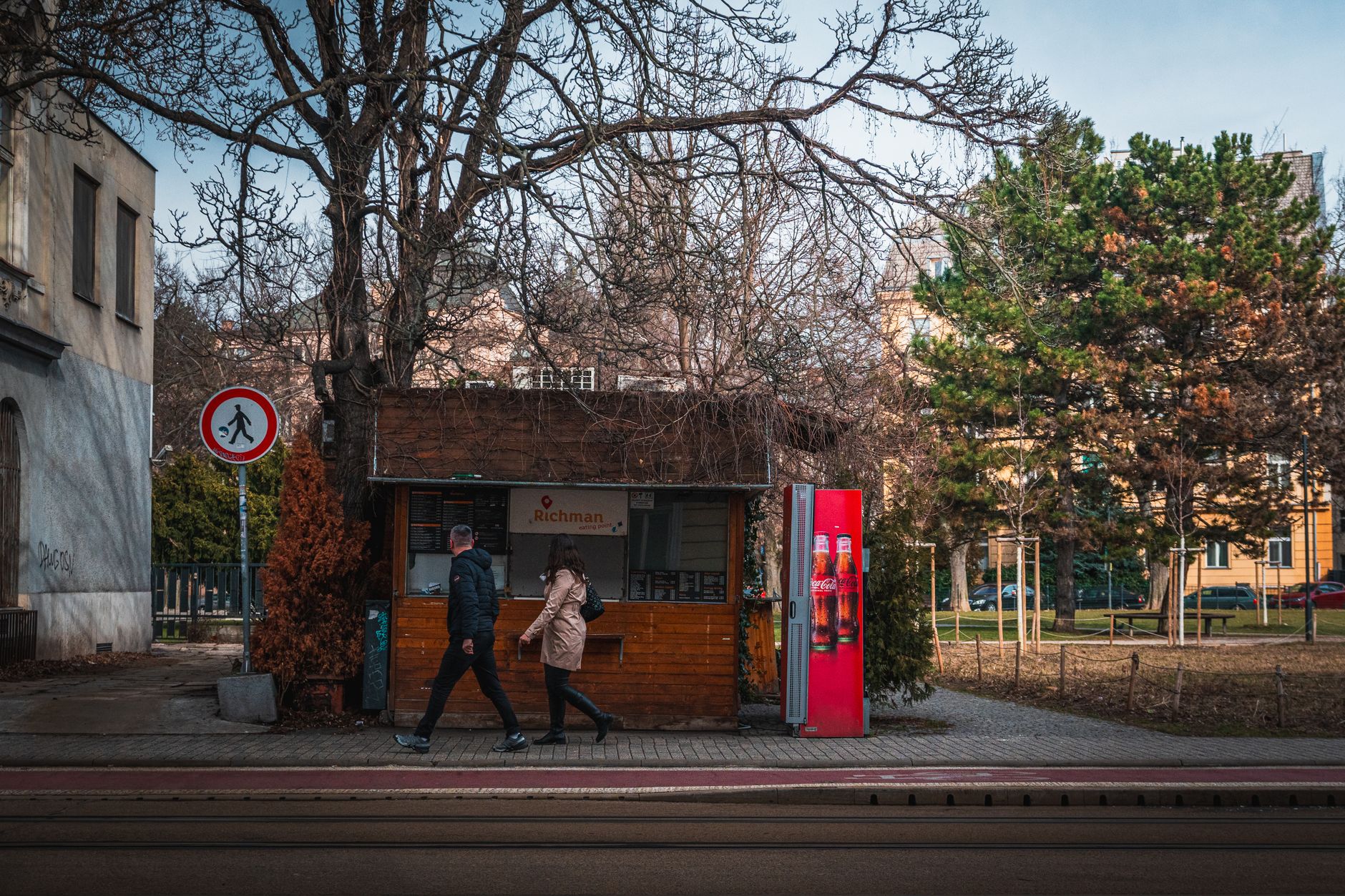 City street scene featuring pedestrians walking past a kiosk with bare trees in winter.