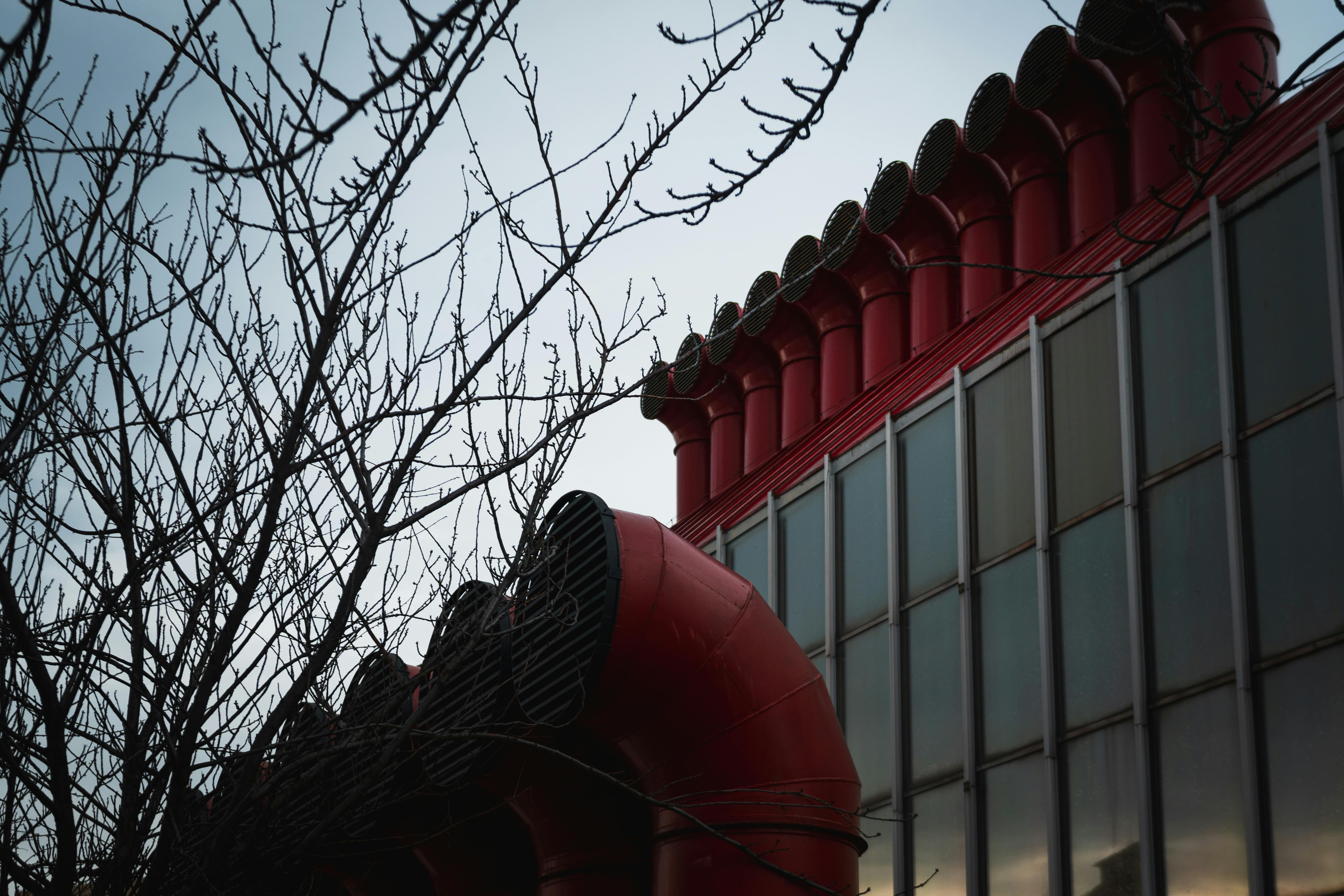 Gratis Sebuah bangunan industri modern dengan ventilasi merah yang khas dan ranting pohon yang gundul di musim dingin. Foto Stok