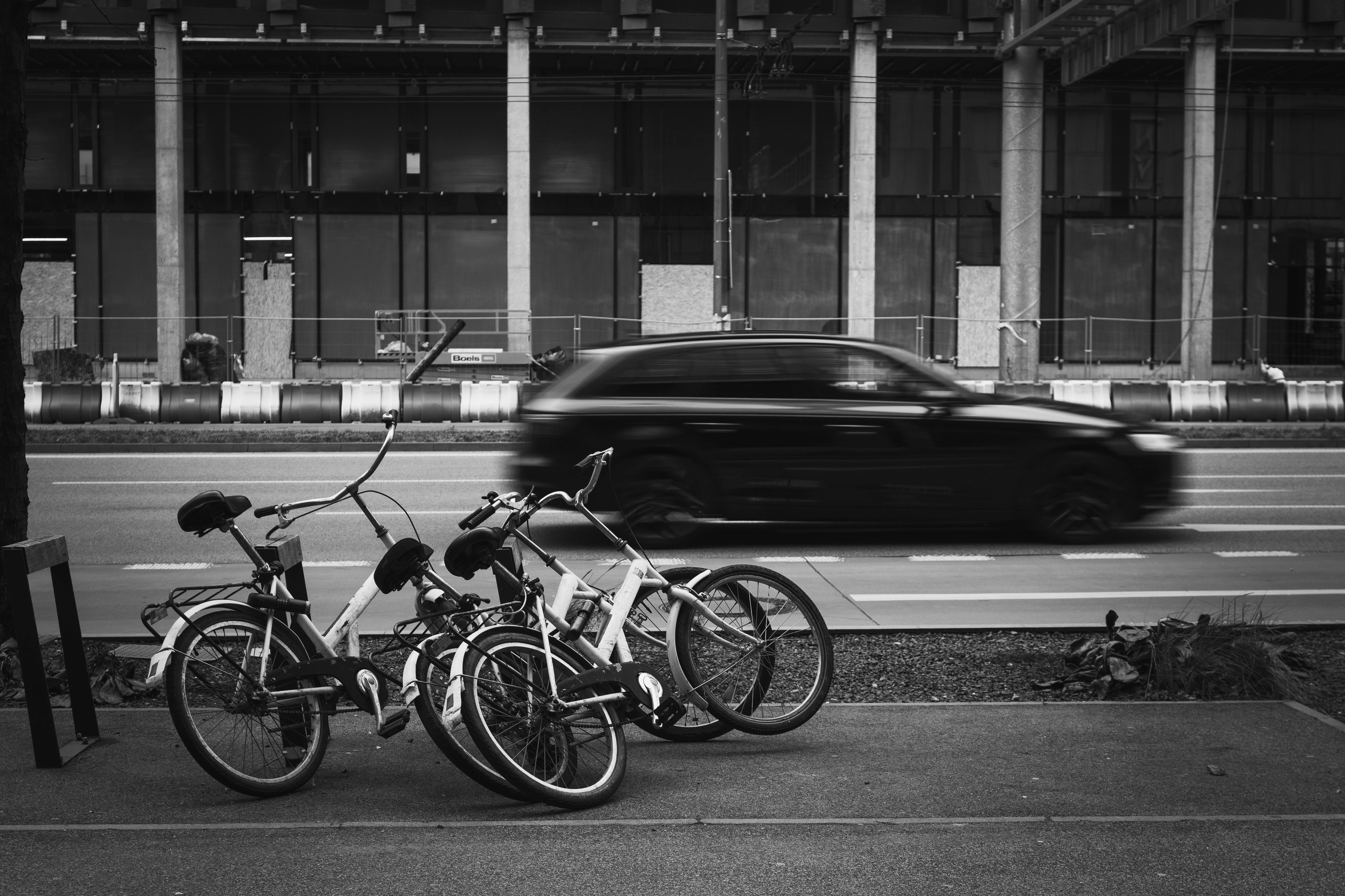 ฟรี ภาพถ่ายขาวดำในเมืองที่มีจักรยานจอดอยู่และรถยนต์กำลังเคลื่อนที่ แสดงให้เห็นถึงความแตกต่างของชีวิตในเมือง คลังภาพถ่าย
