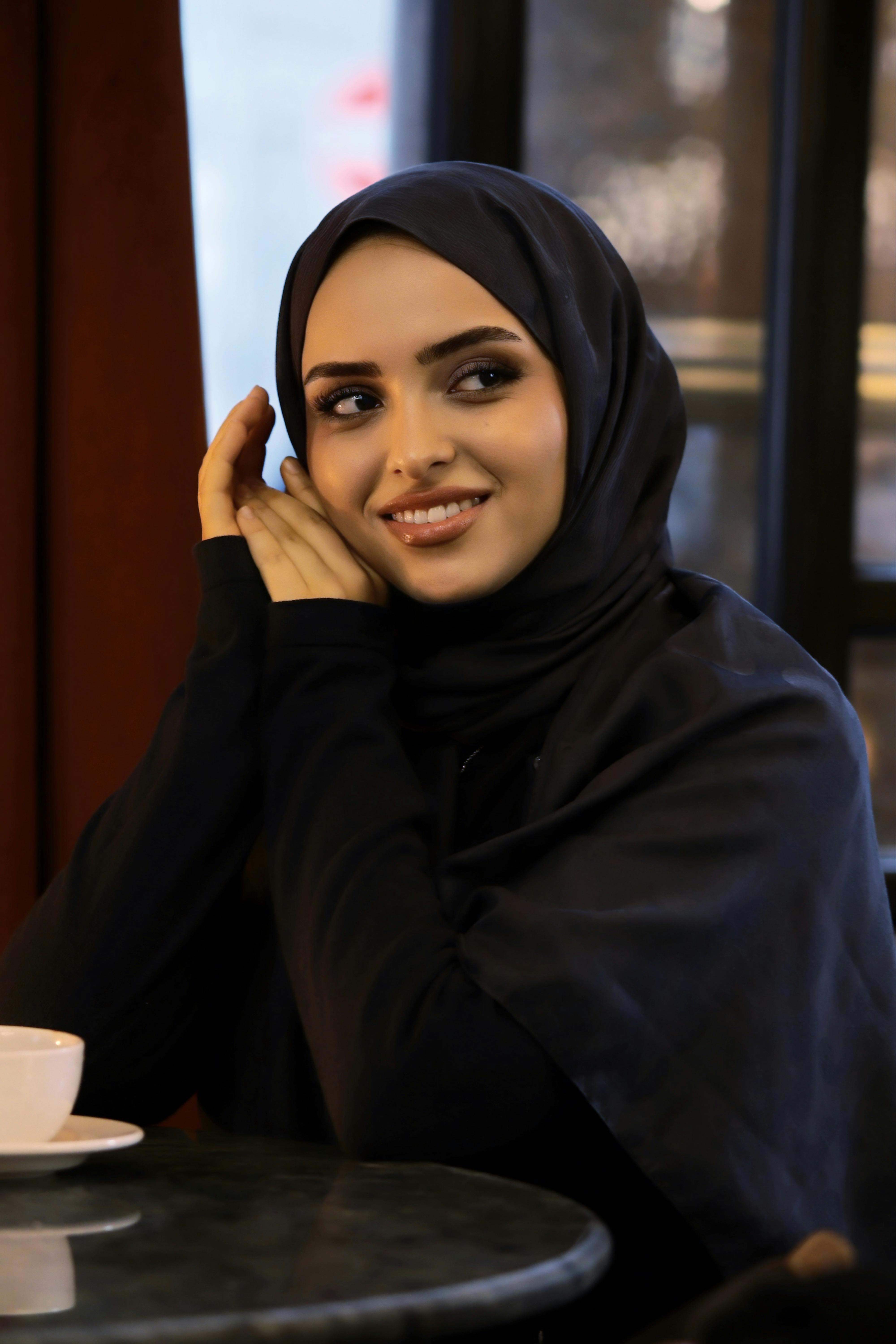 Gratis Una donna che indossa l'hijab sorride calorosamente mentre è seduta al tavolo di un bar, creando un'atmosfera serena e accogliente. Foto a disposizione