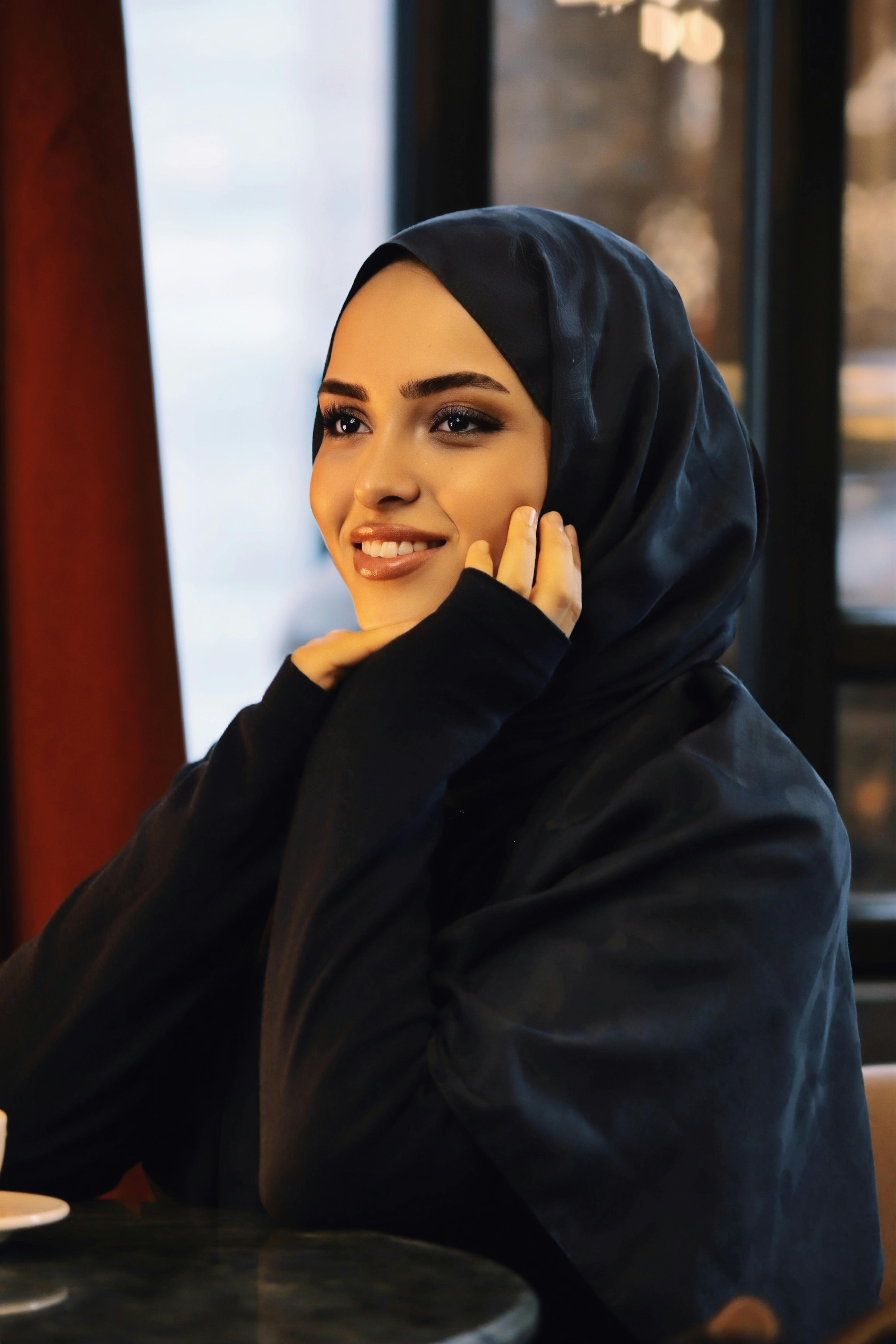 ฟรี หญิงสาวสวมฮิญาบยิ้มอย่างอบอุ่นขณะนั่งอยู่ภายในบ้านริมหน้าต่าง คลังภาพถ่าย