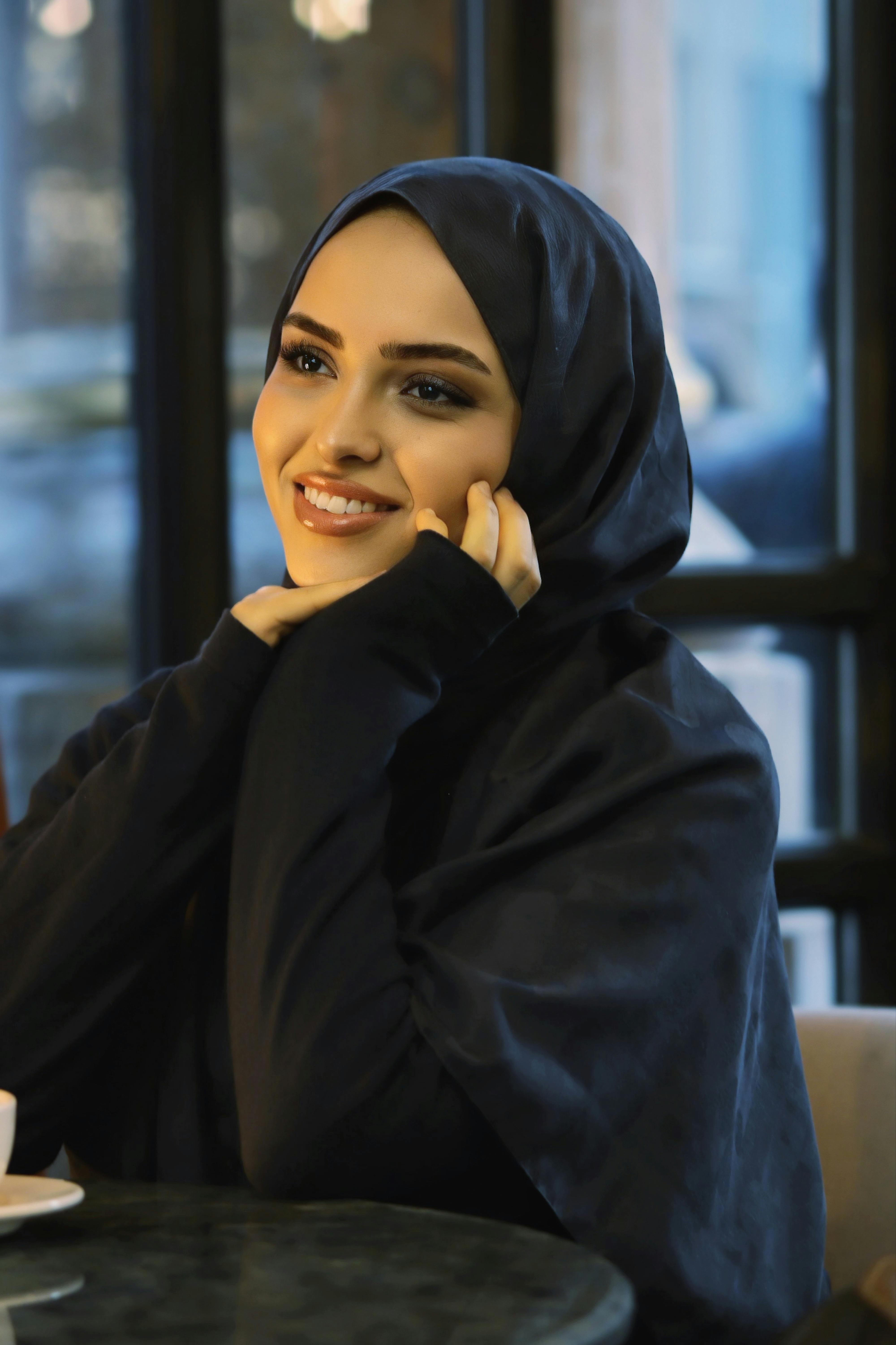 ฟรี หญิงสาวสวมฮิญาบยิ้มอย่างอบอุ่นภายในร้านกาแฟ คลังภาพถ่าย