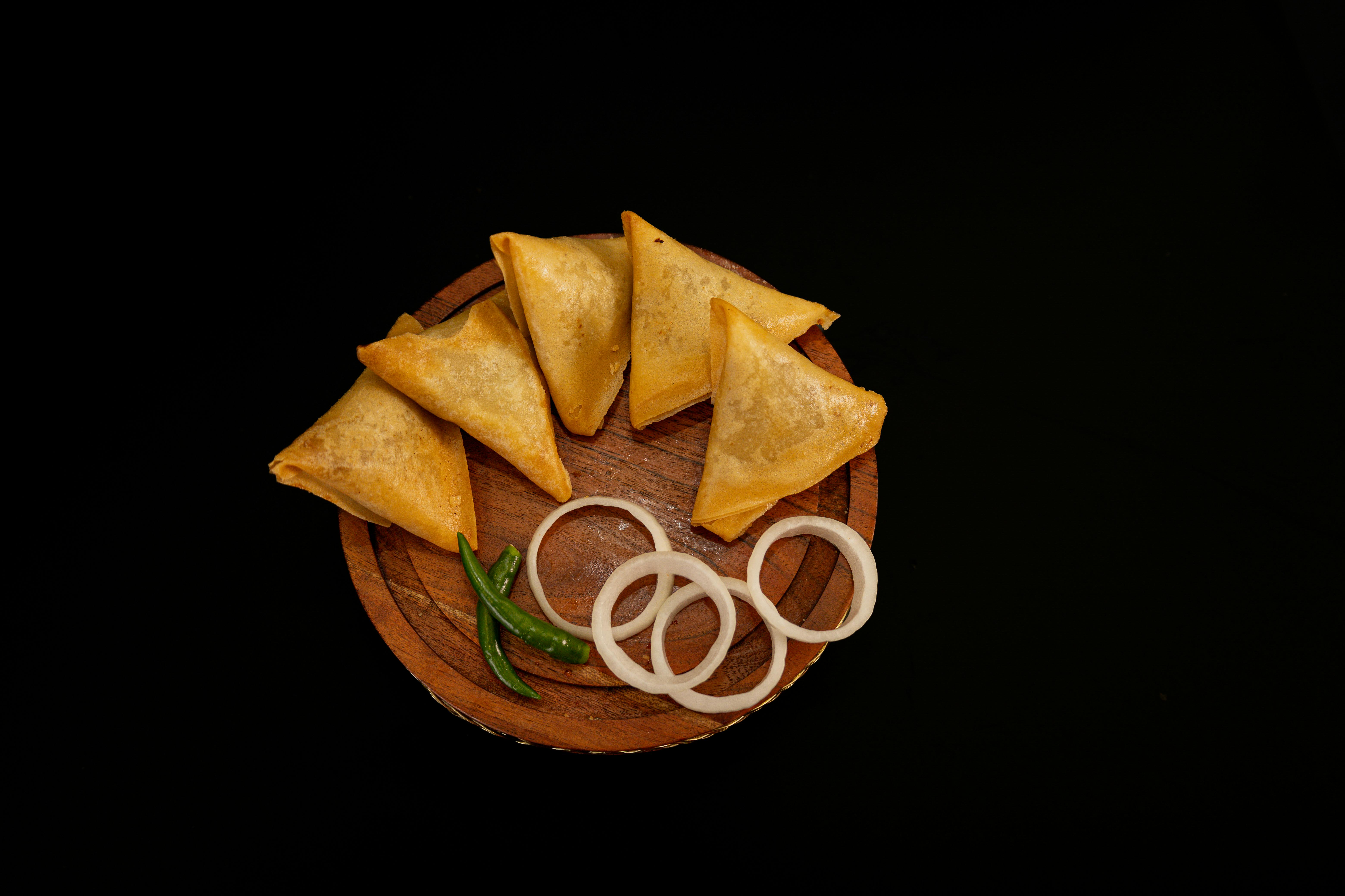 Free Four crispy samosas with onion rings and green chili on a wooden plate against black background. Stock Photo