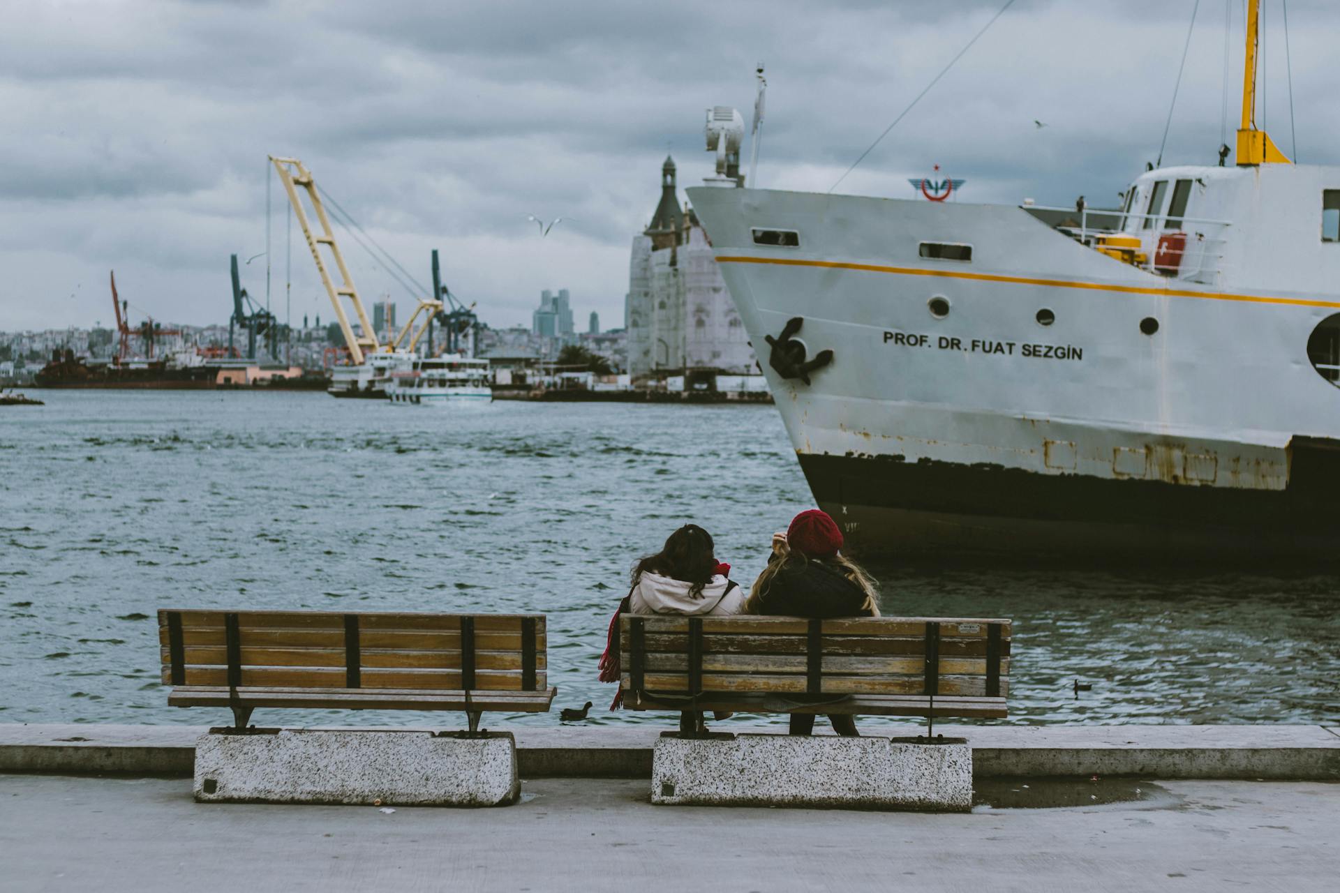 Two women sitting on a bench near a harbor while a large ship passes by.