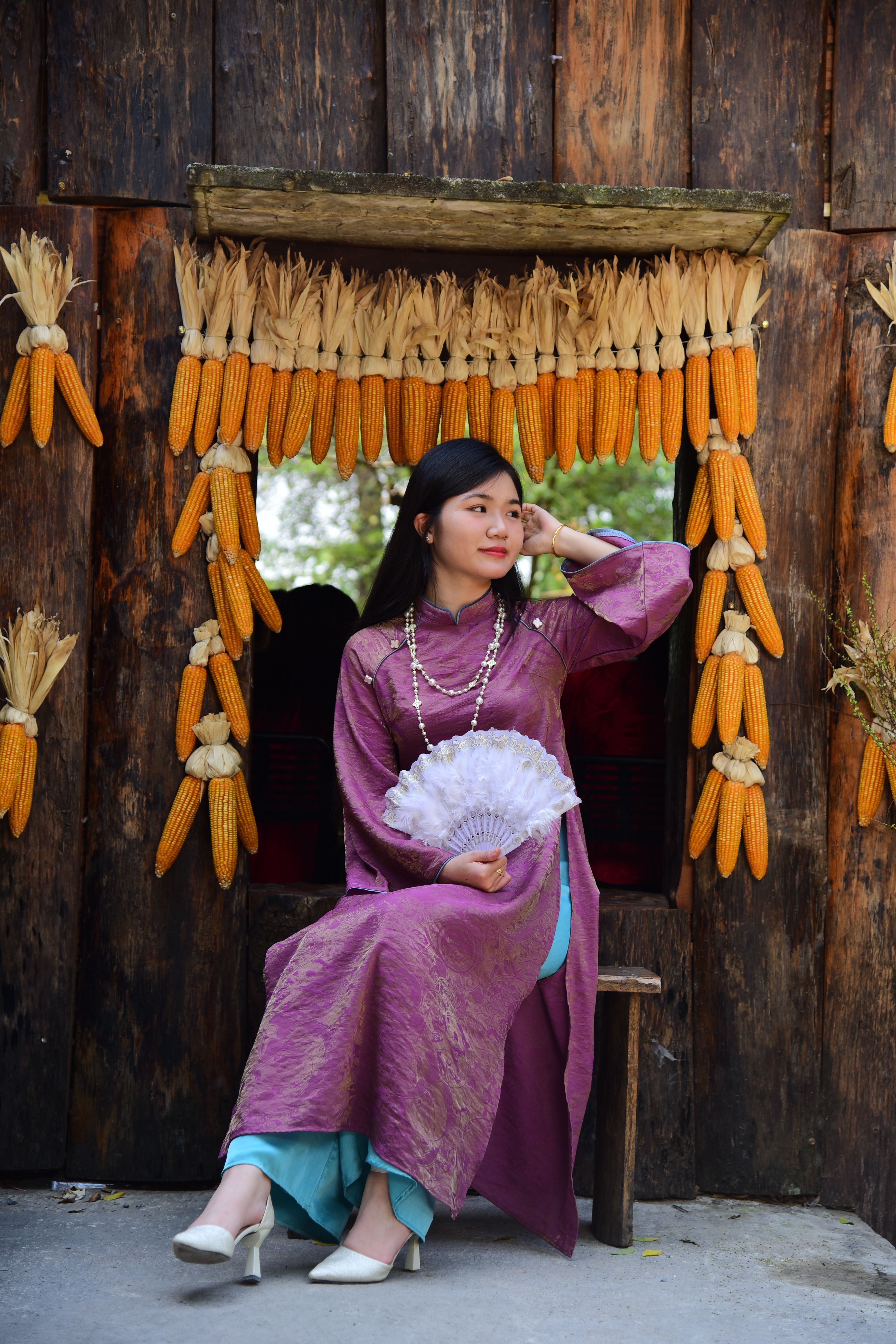 Woman in traditional attire seated among hanging corn cobs, holding a fan.
