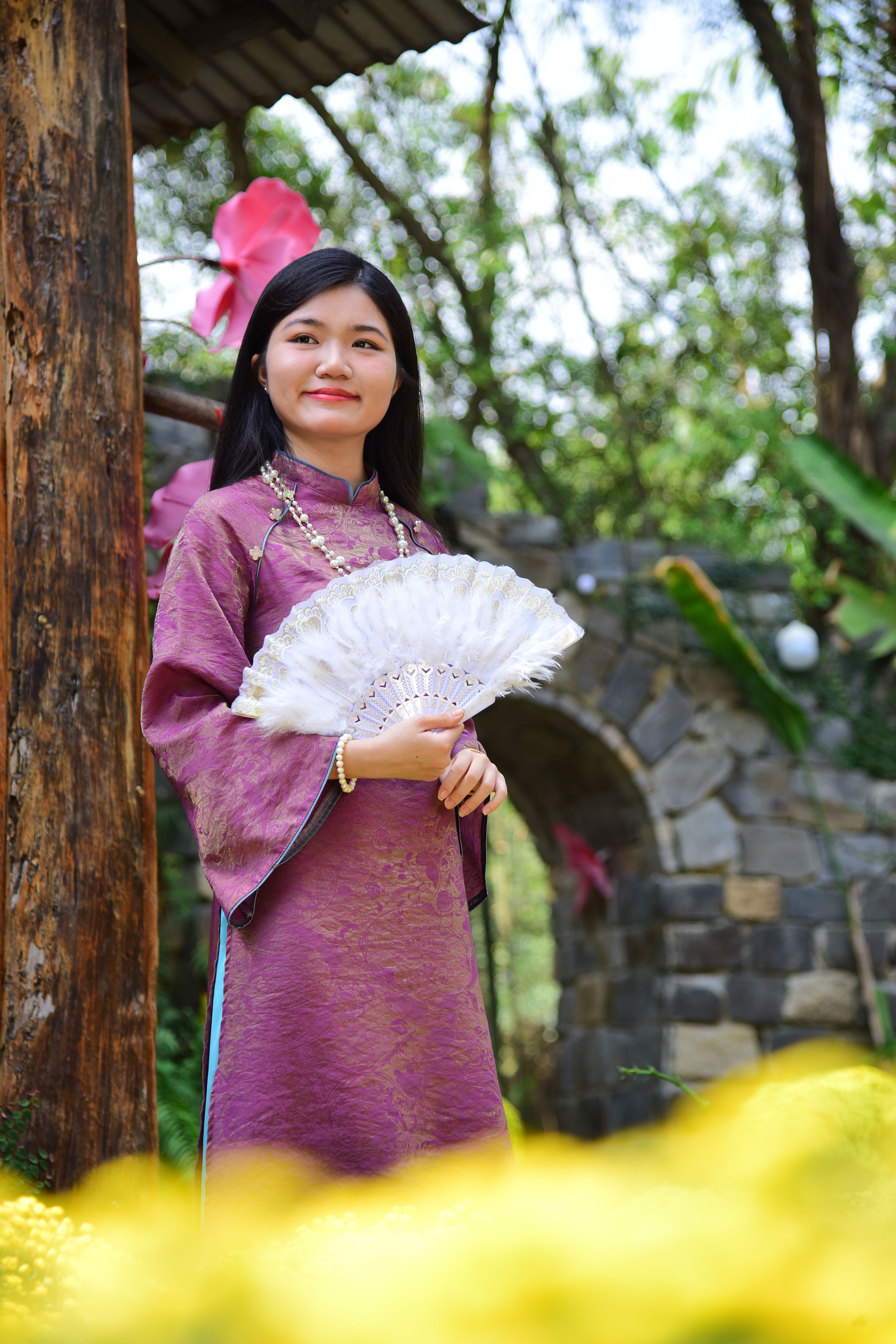 A woman in a purple traditional dress holds a fan in an outdoor garden setting.