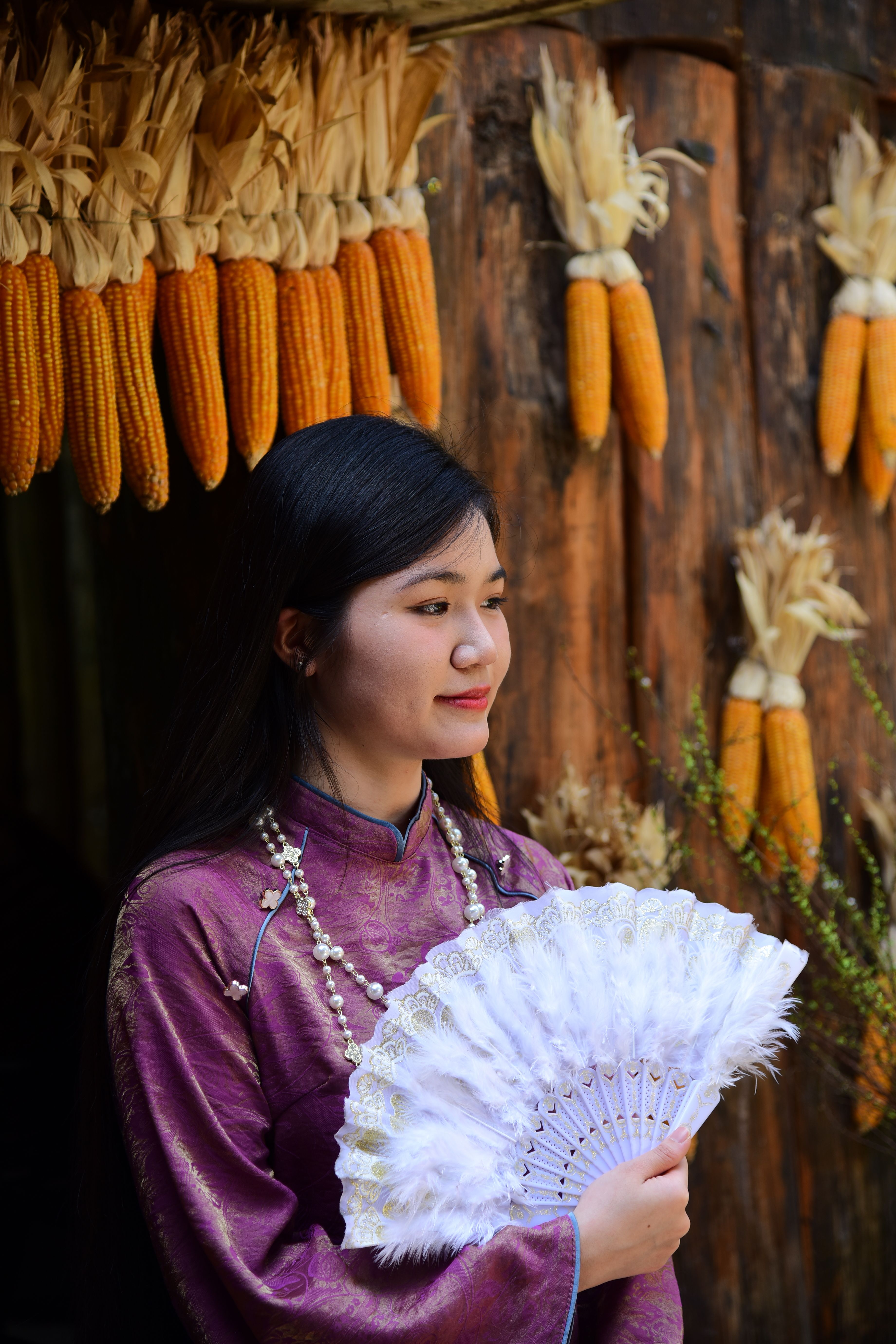 Woman in purple traditional outfit holding a fan, with corn decorations in background.