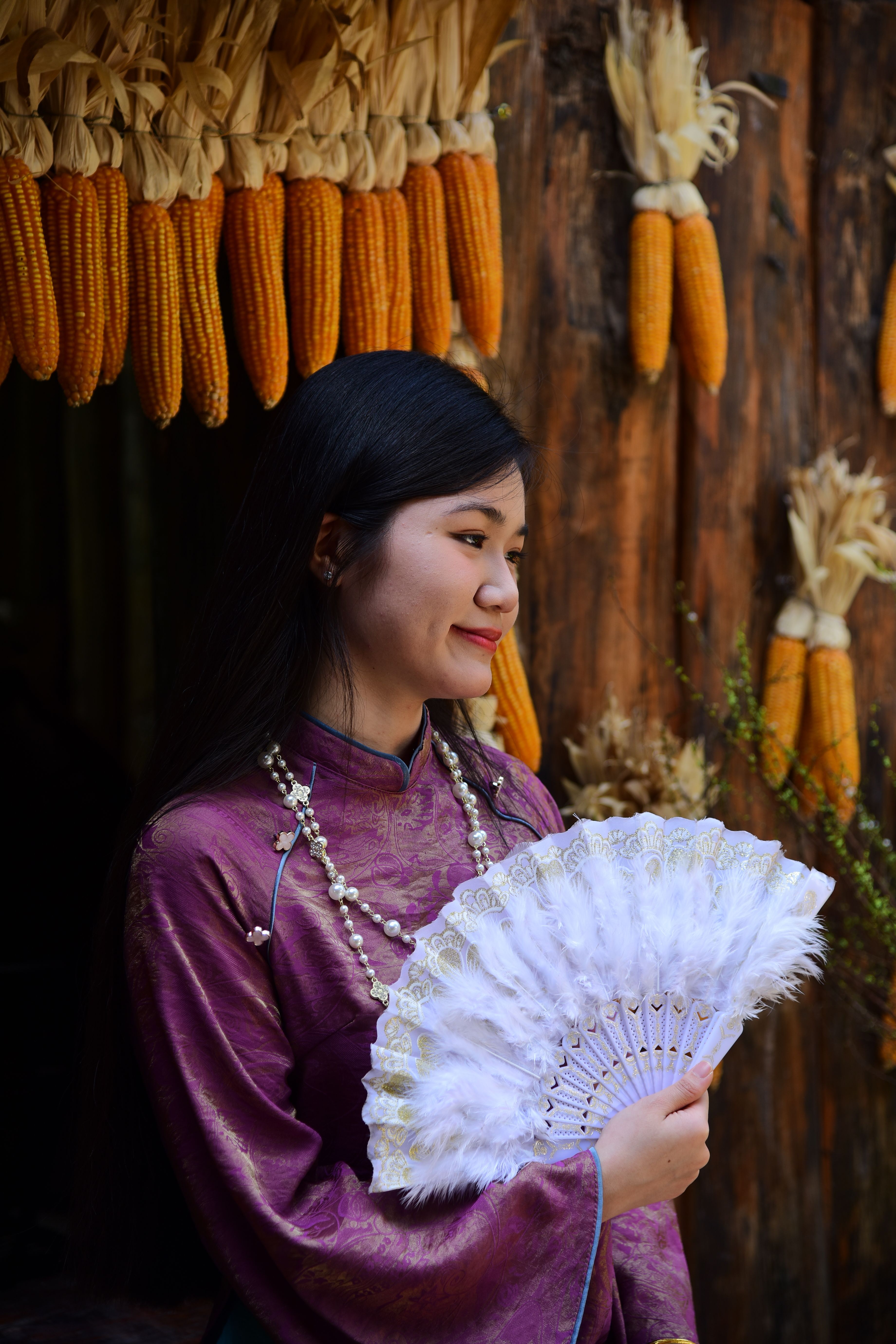 Elegant Asian woman in traditional attire with a fan, surrounded by hanging corn.