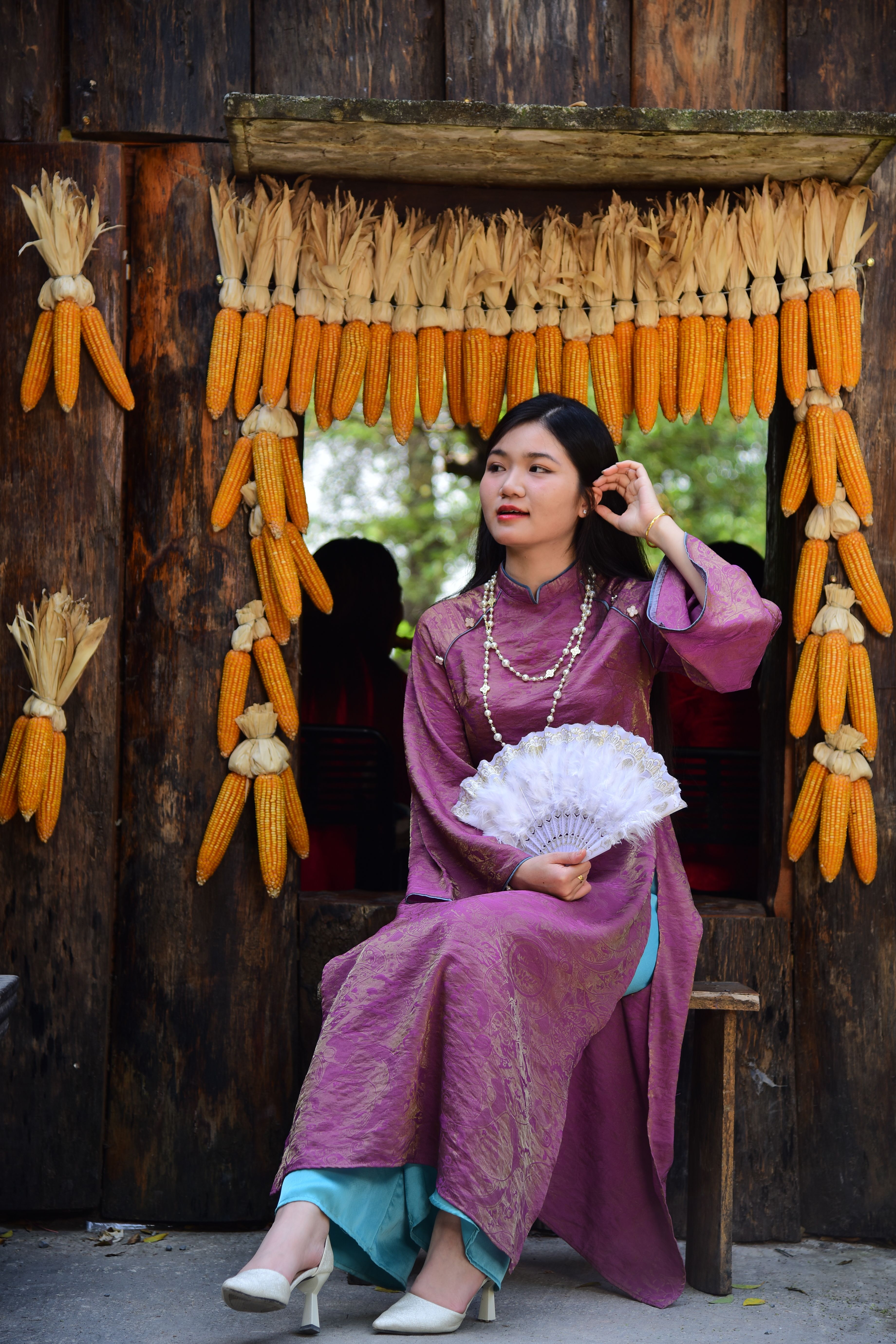 Woman in traditional Vietnamese attire seated near drying corn, exuding cultural elegance.