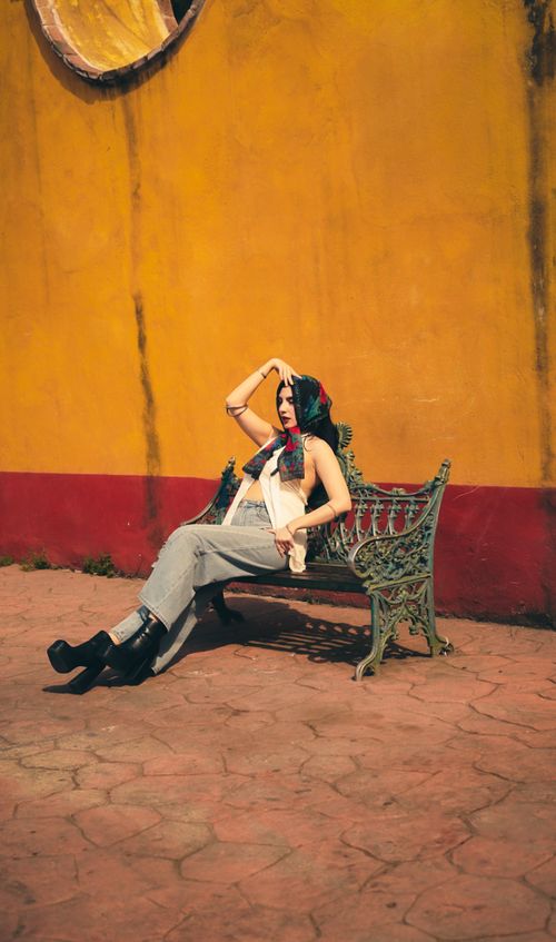 Free Stylish woman posing on an ornate bench against a vibrant yellow wall, exuding elegance and modern fashion. Stock Photo