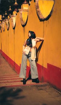 Stylish woman in wide-leg jeans and scarf poses against a colorful wall on a sunny day.