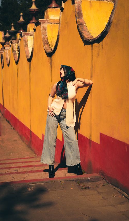 Free Stylish woman in wide-leg jeans and scarf poses against a colorful wall on a sunny day. Stock Photo