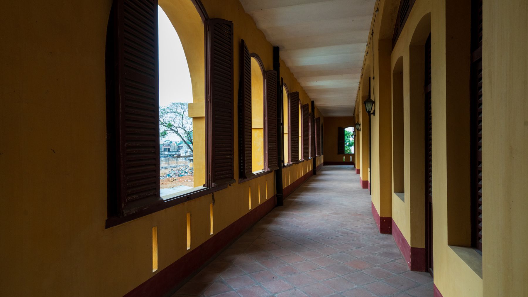 Sunlit corridor in a historic colonial building featuring arched windows and wooden shutters.