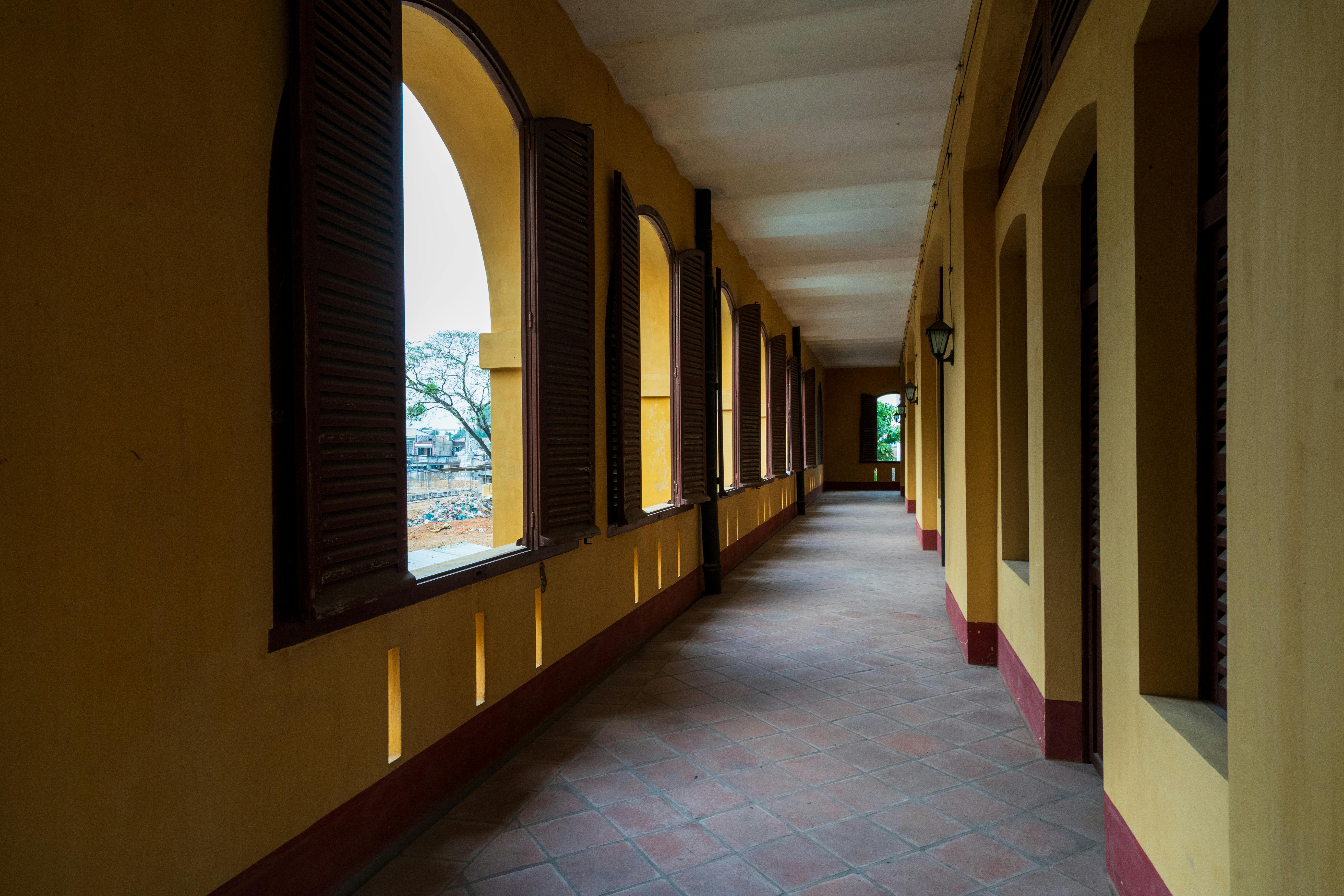 Gratuit Couloir baigné de soleil dans un bâtiment colonial historique, orné de fenêtres cintrées et de volets en bois. Photos