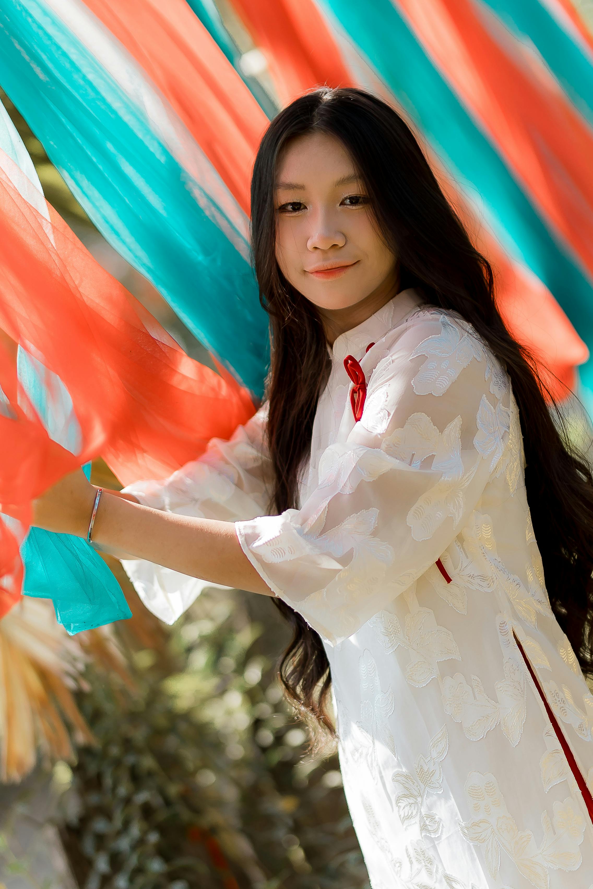 Gratis Una giovane donna in abito tradizionale bianco gioca con tessuti colorati e fluenti in un ambiente esterno. Foto a disposizione