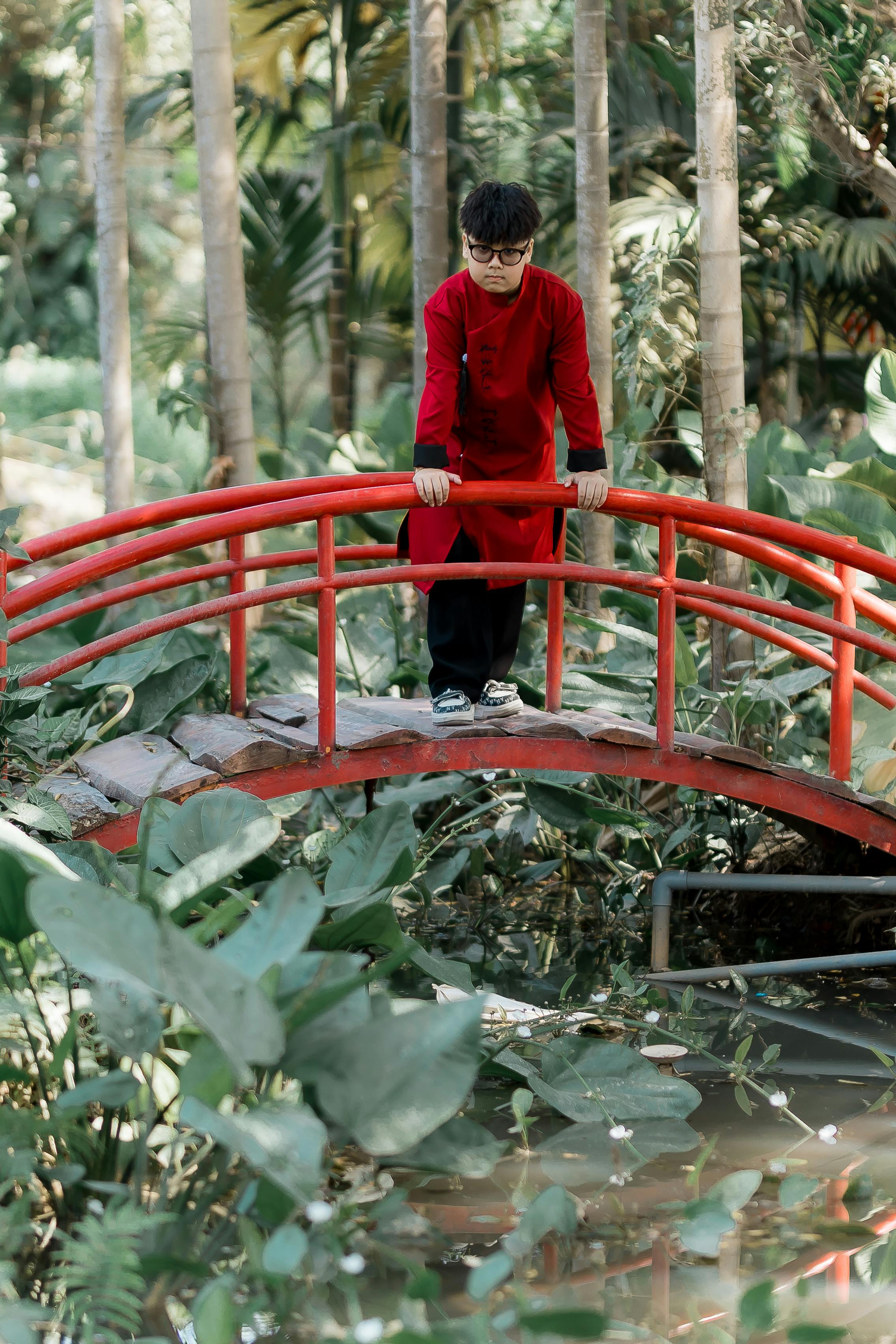 bezplatná Muž v tradičním červeném oděvu stojí na malebném červeném mostě uprostřed bujné zahrady. Základová fotografie