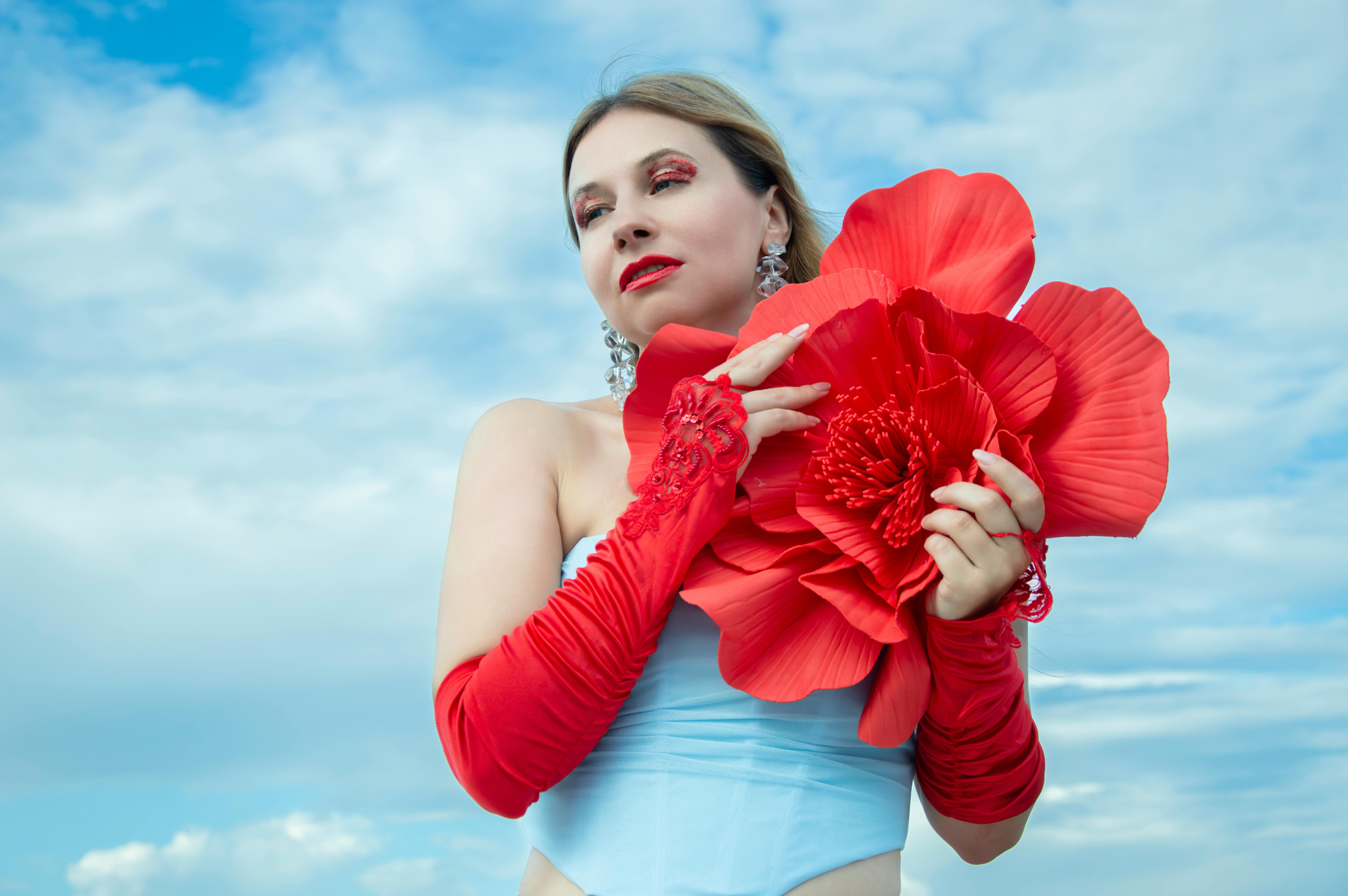 Gratis Una donna elegante che tiene in mano un enorme fiore rosso contro un cielo azzurro terso, esprimendo stile ed eleganza. Foto a disposizione