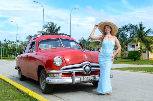 Stylish woman in blue dress and straw hat stands by a vintage red car on a sunny day.