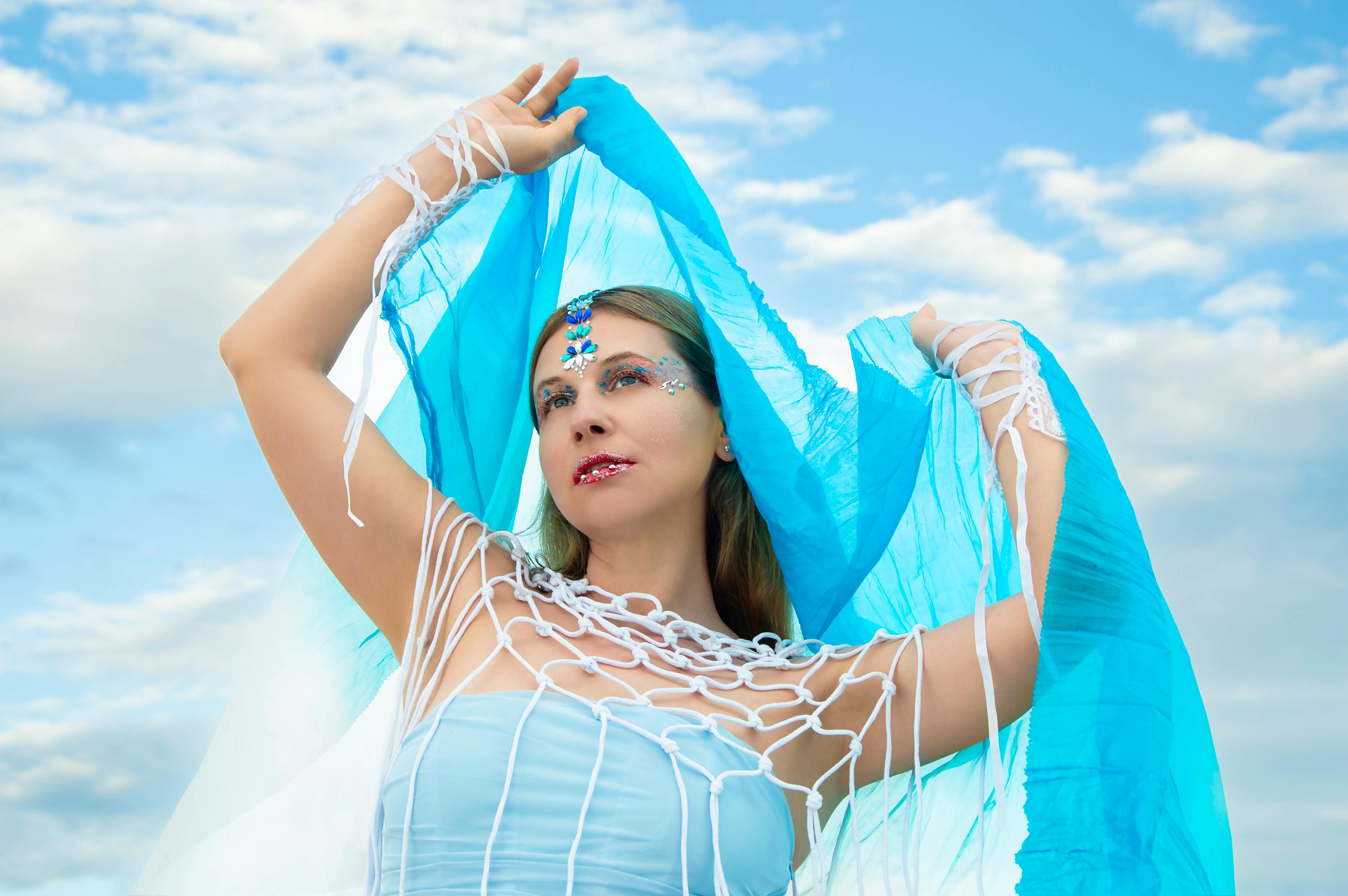 Free A woman posing outdoors with blue fabric and artistic face makeup under a bright sky. Stock Photo