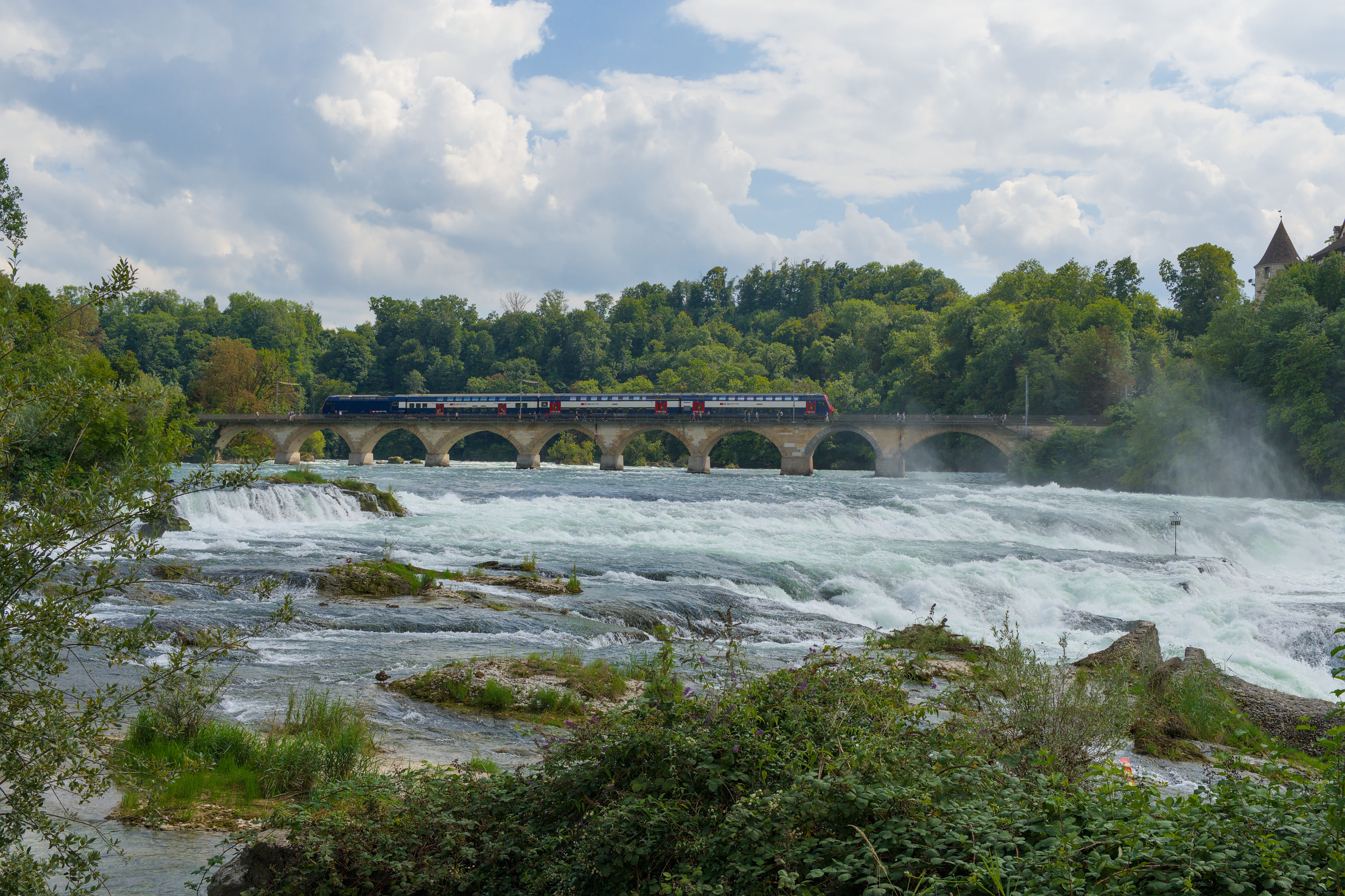 Breathtaking view of Rhine Falls with a train crossing a historic bridge, lush greenery and blue skies. - Schaffhausen