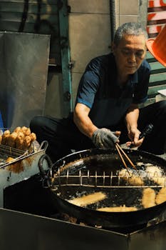 An adult man fries food in a large pan at an outdoor night market. Urban ambiance.