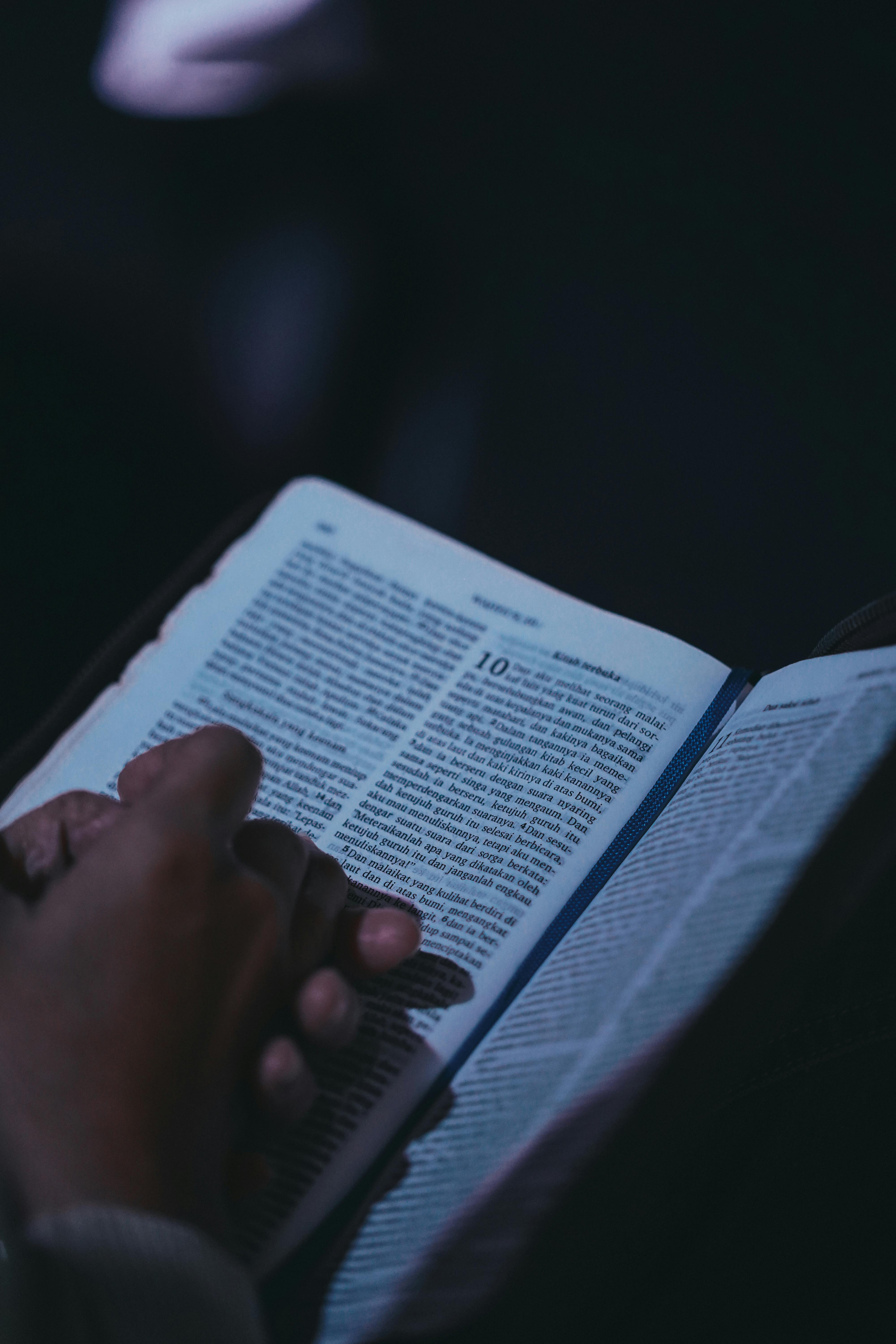 ฟรี ภาพระยะใกล้ของมือที่กำลังถือหนังสือเปิดอยู่ ราวกับกำลังครุ่นคิด ในสภาพแวดล้อมที่มีแสงสลัว คลังภาพถ่าย