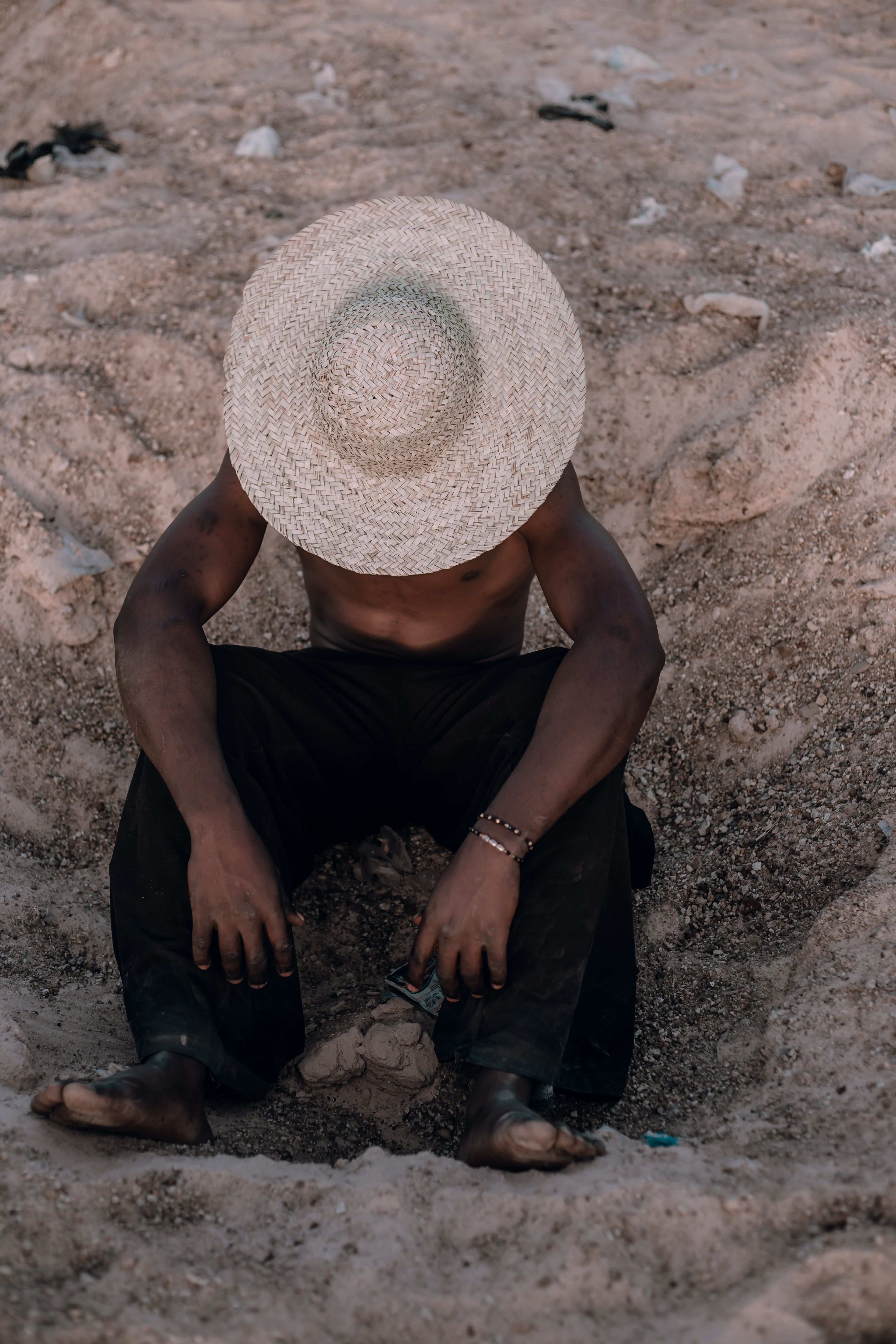 A solitary farmer sits resting in a sandy area, wearing a wide-brimmed straw hat, in Nigeria's rural KT region.