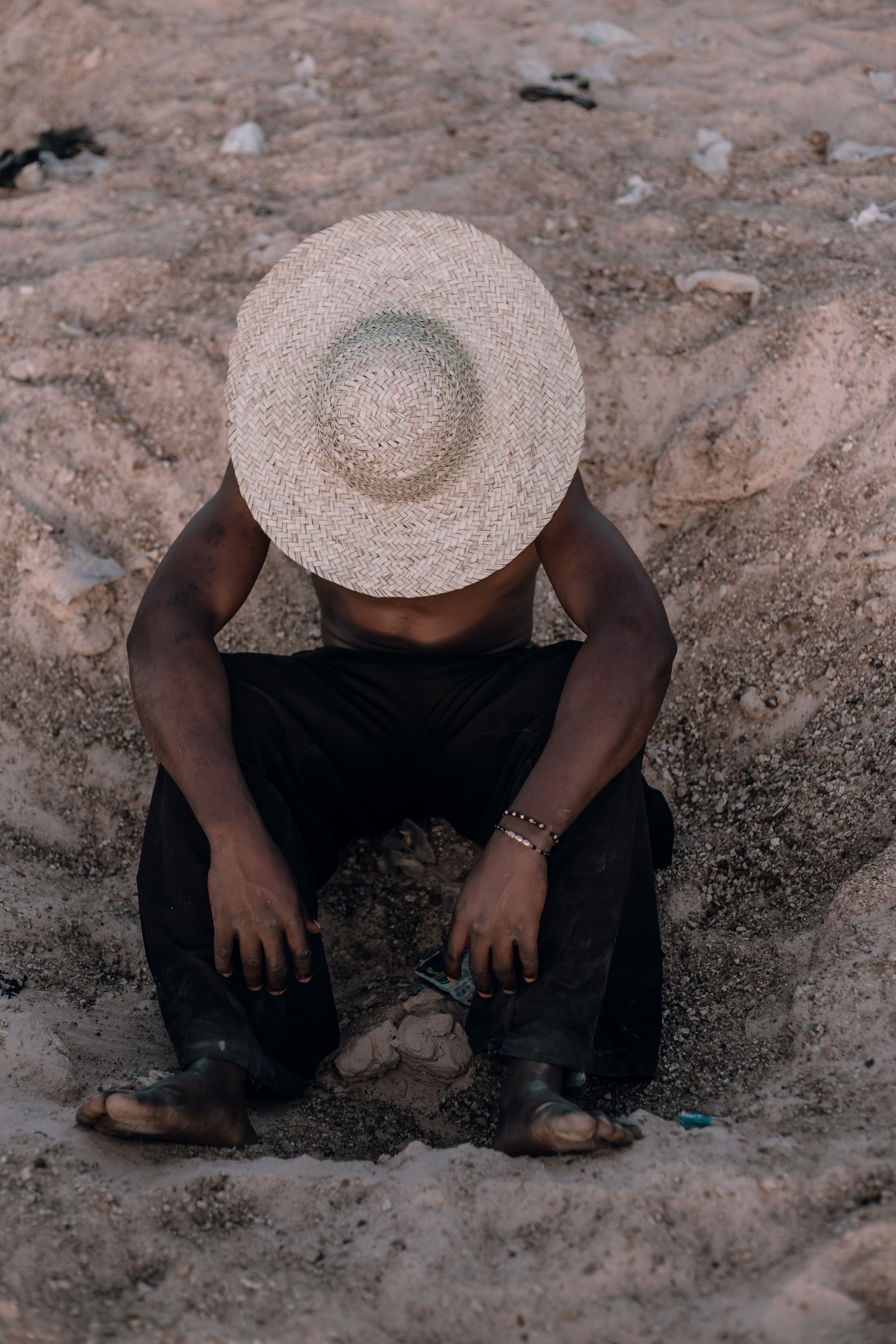 Man with straw hat sits contemplatively on a Nigerian beach, sun casting shadows, creating a peaceful scene.