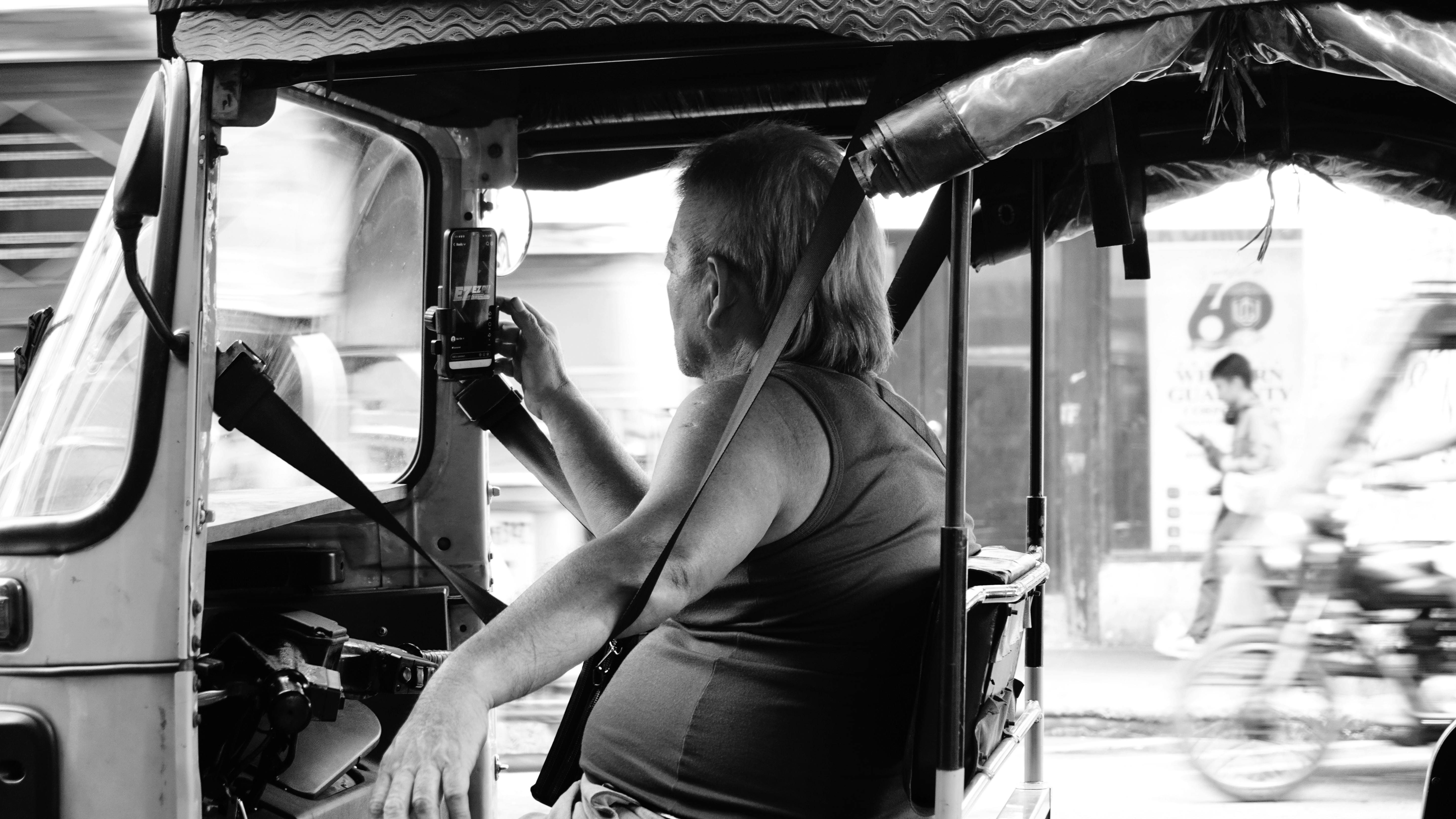 Free Black and white photo of a man operating a tuk-tuk and using a smartphone on a bustling street. Stock Photo