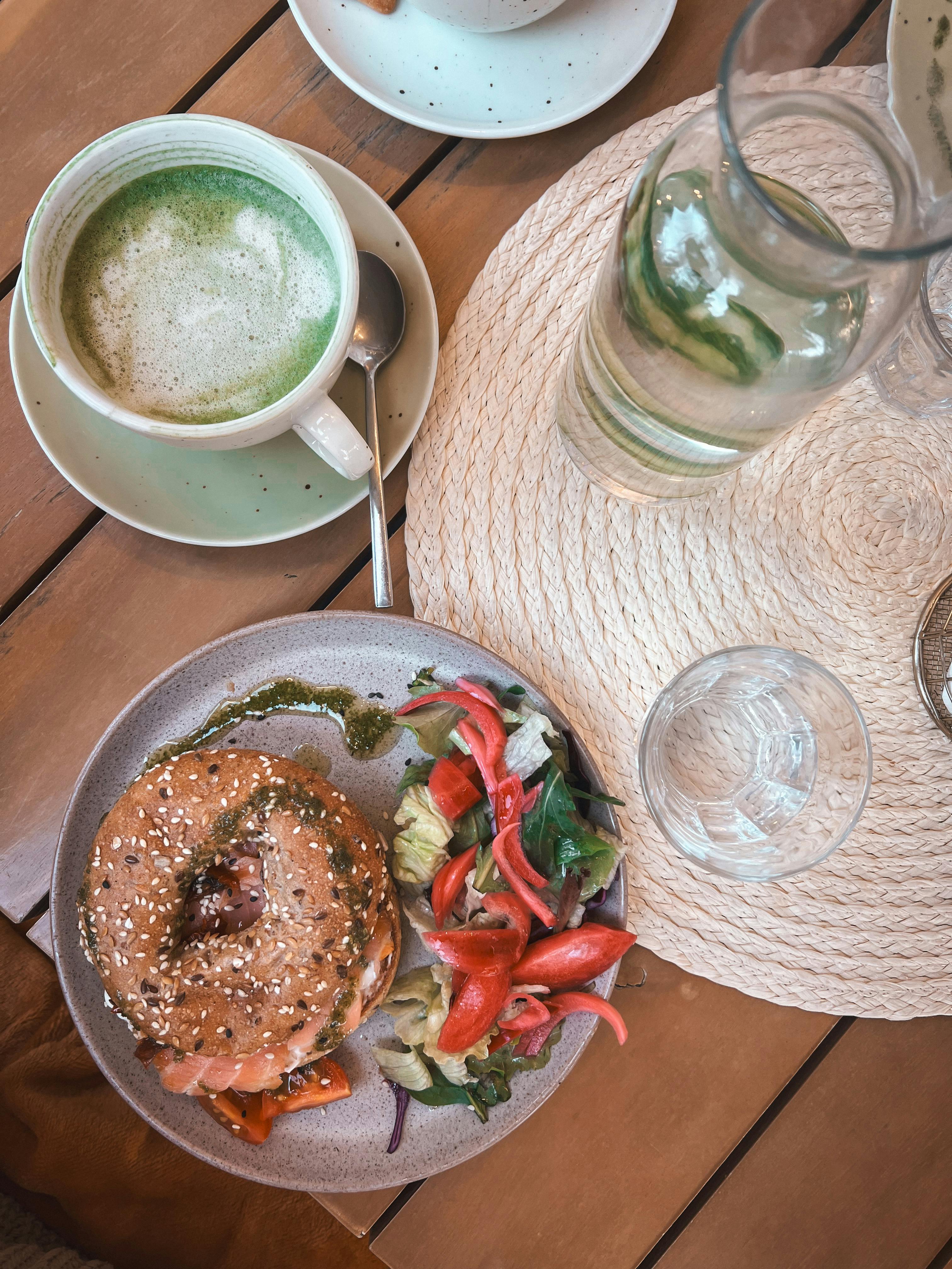 ฟรี ภาพมุมสูงของอาหารเช้าเพื่อสุขภาพ ประกอบด้วยเบเกิล สลัด และชาเขียวมัทฉะ วางอยู่บนโต๊ะไม้ คลังภาพถ่าย