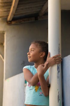 A young girl looking thoughtfully outside while leaning on a post.