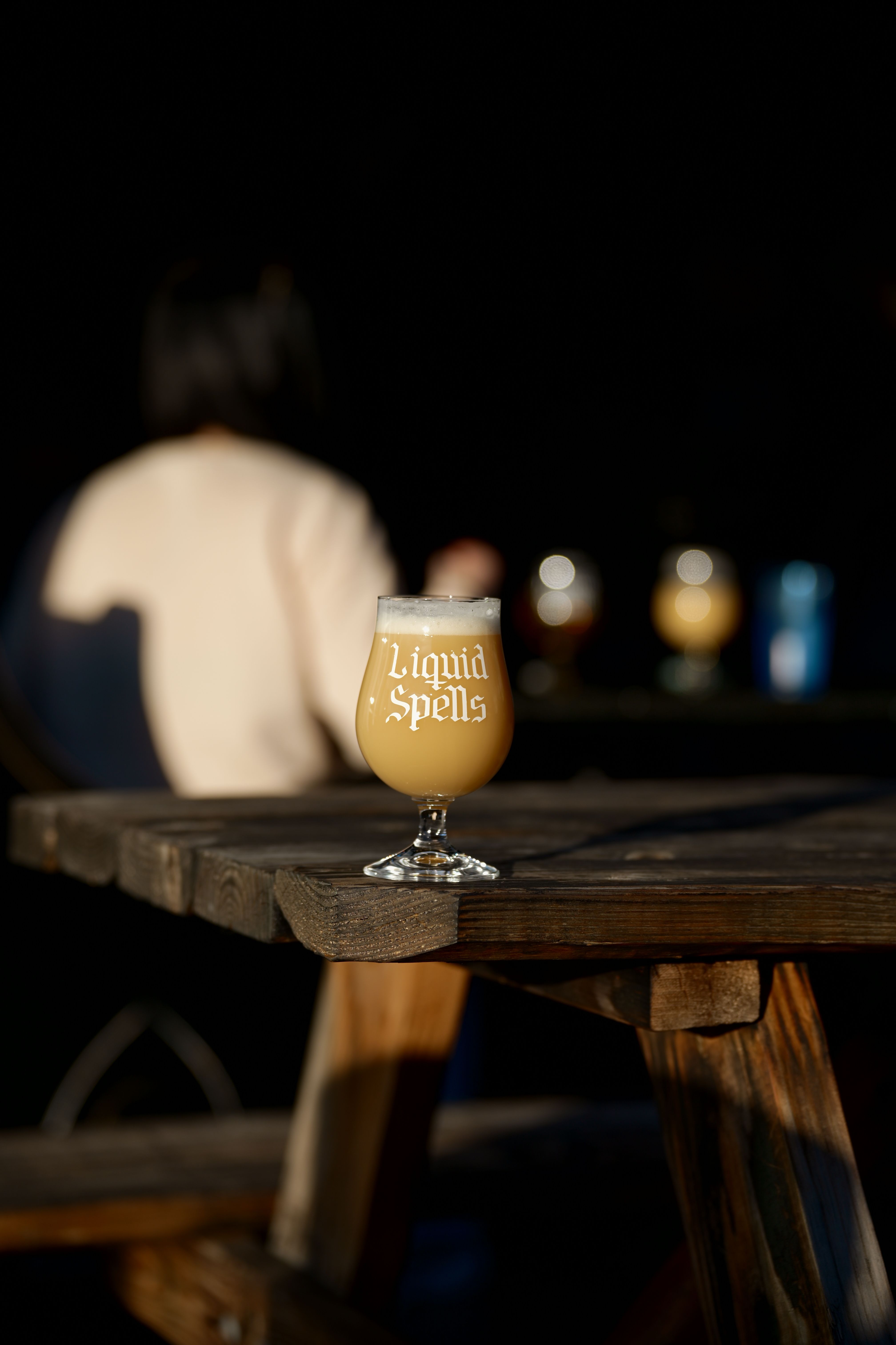 A glass of 'Liquid Spells' craft beer sits on a rustic wooden table outdoors with people in the background.