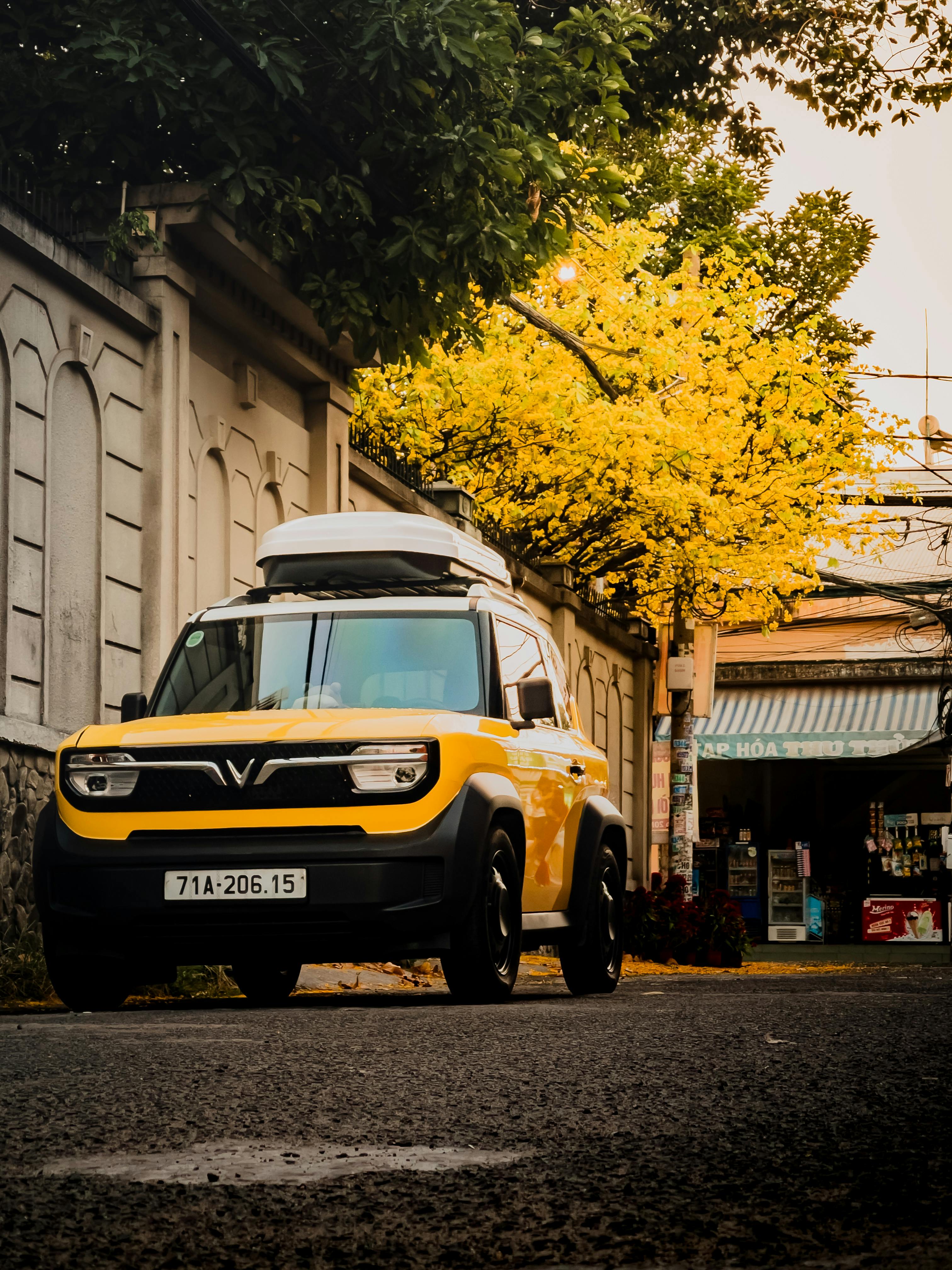 Ilmainen Kirkkaankeltainen katumaasturi pysäköitynä puun alle, jonka lehdet ovat kirkkaankeltaisia, vietnamilaisella kadulla syksyllä. Kuvapankkikuva