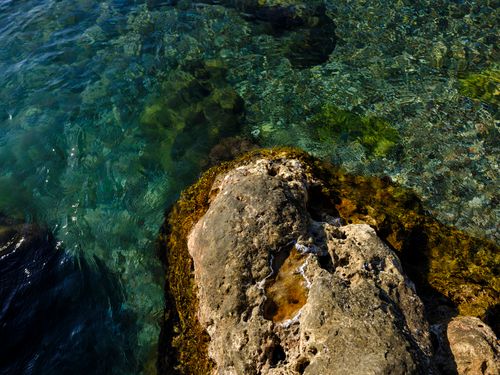 Free A tranquil scene of clear blue waters revealing rocks beneath, alongside a coastal rock outcrop. Stock Photo