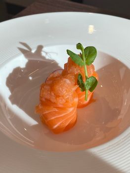 Fresh salmon sashimi garnished with microgreens on a white plate under sunlight.