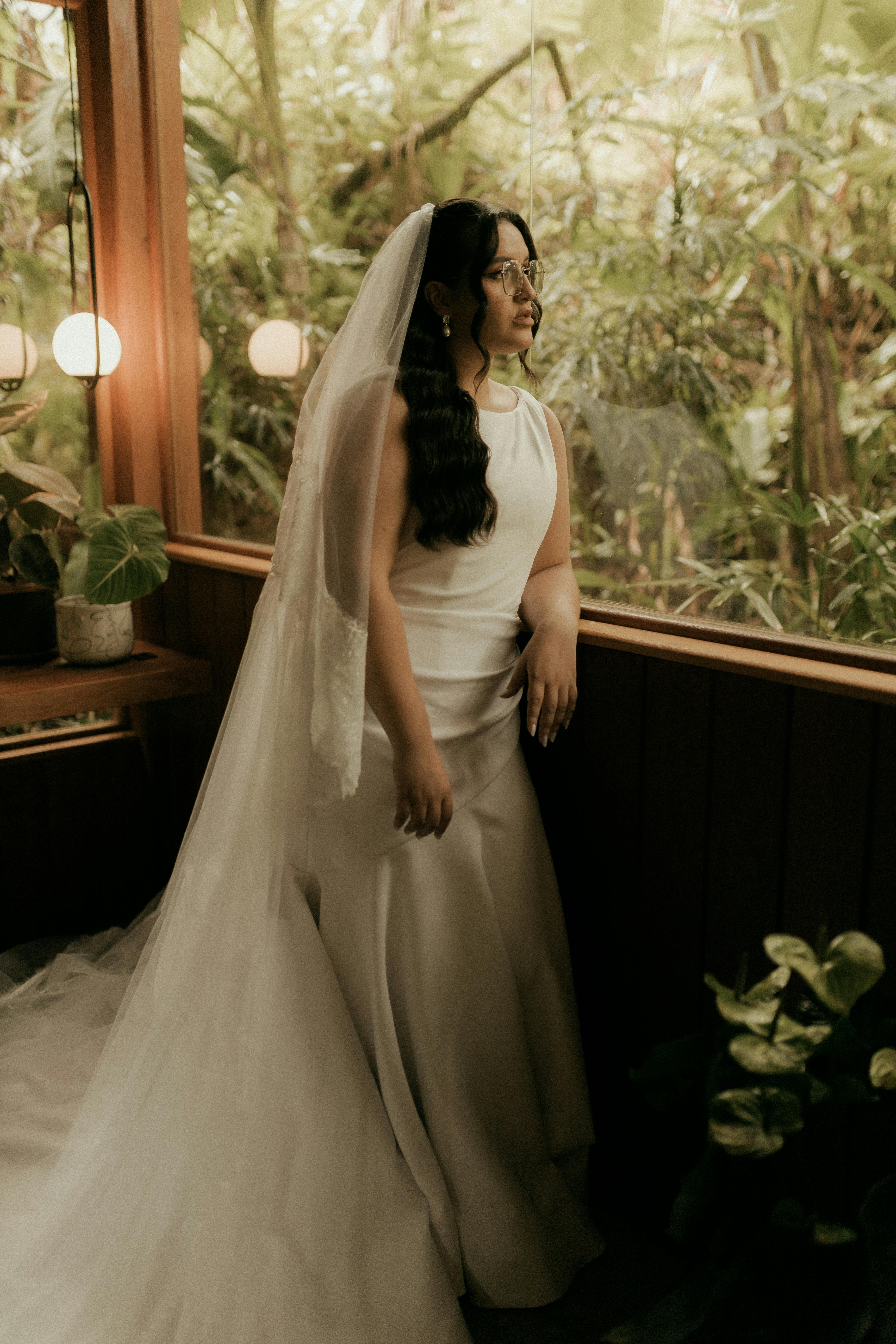 ฟรี เจ้าสาวยืนอย่างสง่างามอยู่ภายในบ้านริมหน้าต่าง ล้อมรอบด้วยต้นไม้และแสงไฟอ่อนๆ คลังภาพถ่าย
