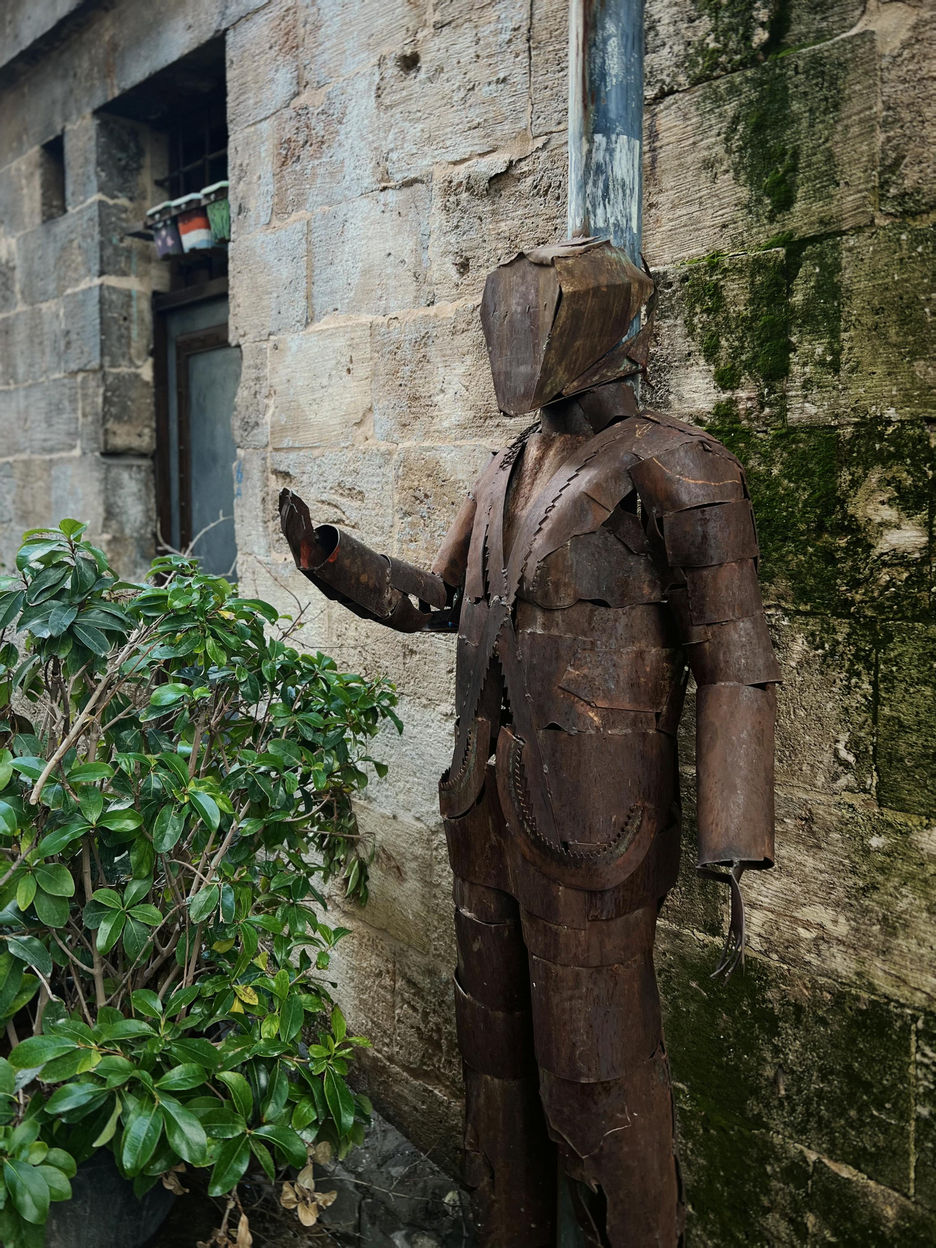 bezplatná Rustikální kovová socha vhodné postavy vedle staré kamenné budovy venku. Základová fotografie