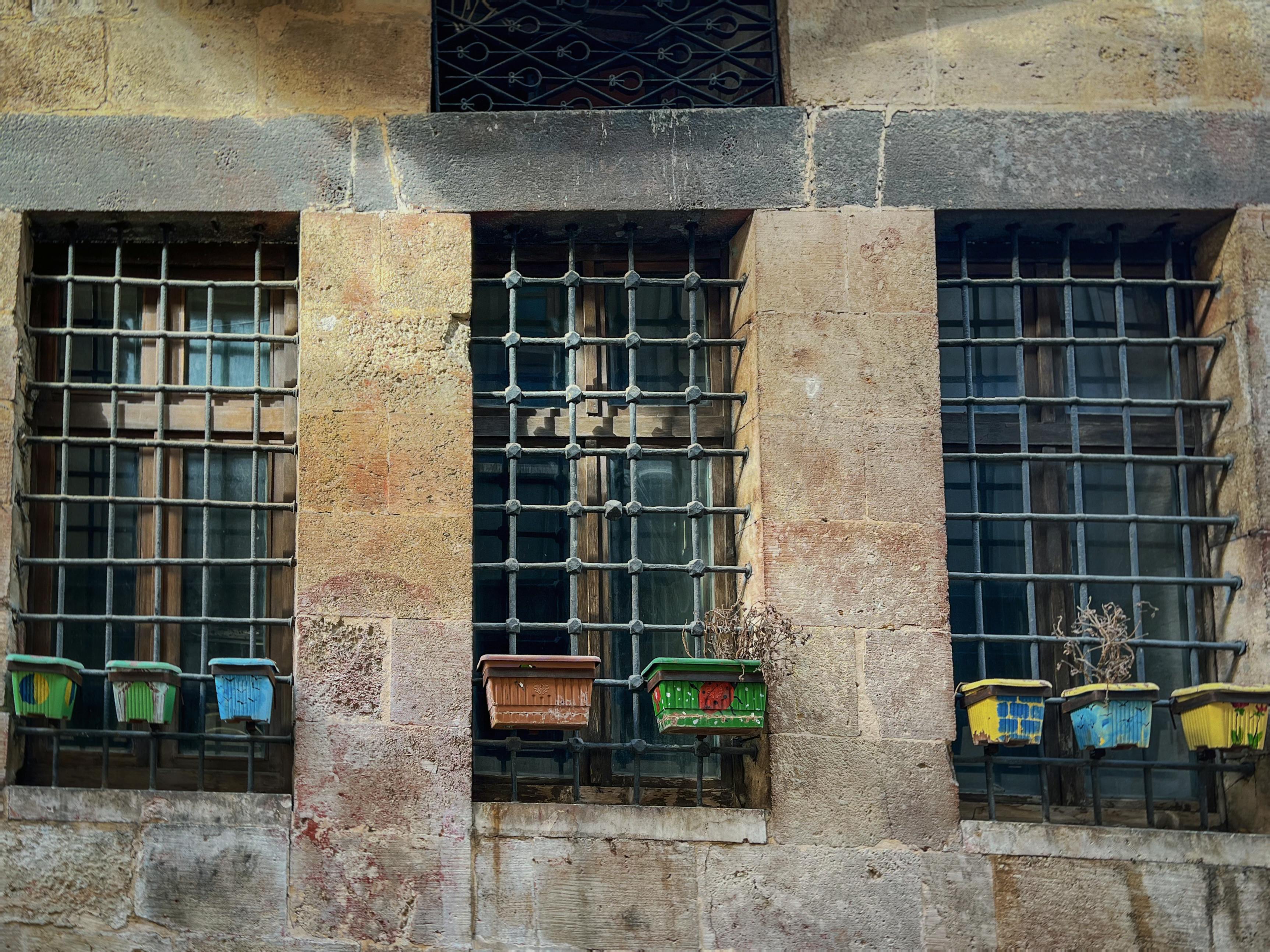 Gratuit Trois fenêtres anciennes à grilles en fer forgé et des pots de fleurs colorés sur un mur de pierre rustique. Photos