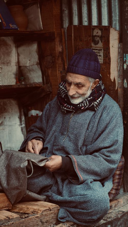 Free Senior man in Srinagar, Kashmir sewing with traditional attire, captured indoors in a warm, muted setting. Stock Photo