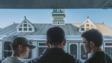 Three young adults converse at a train station, with urban architecture in the background.