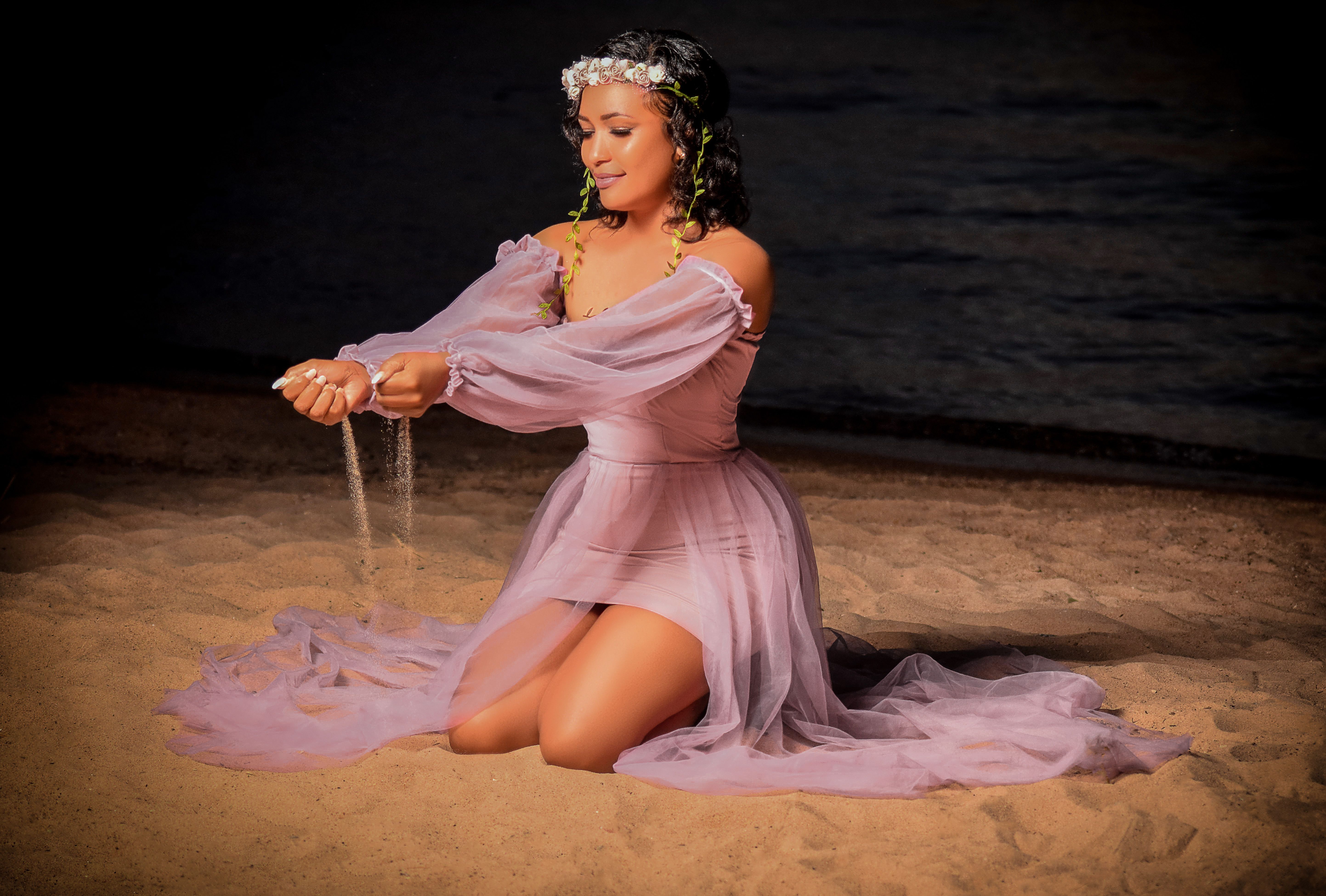 A woman in a flowing dress pours sand on a beach during a serene night.