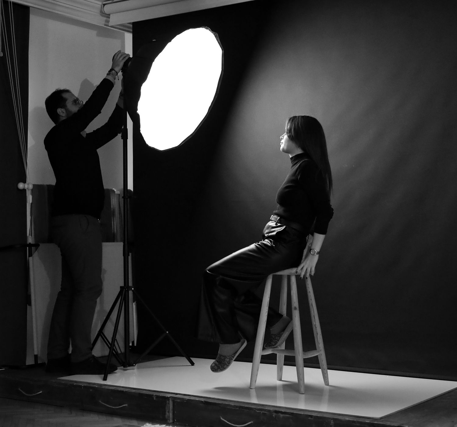 A photographer adjusts studio lighting for a female model seated on a stool.