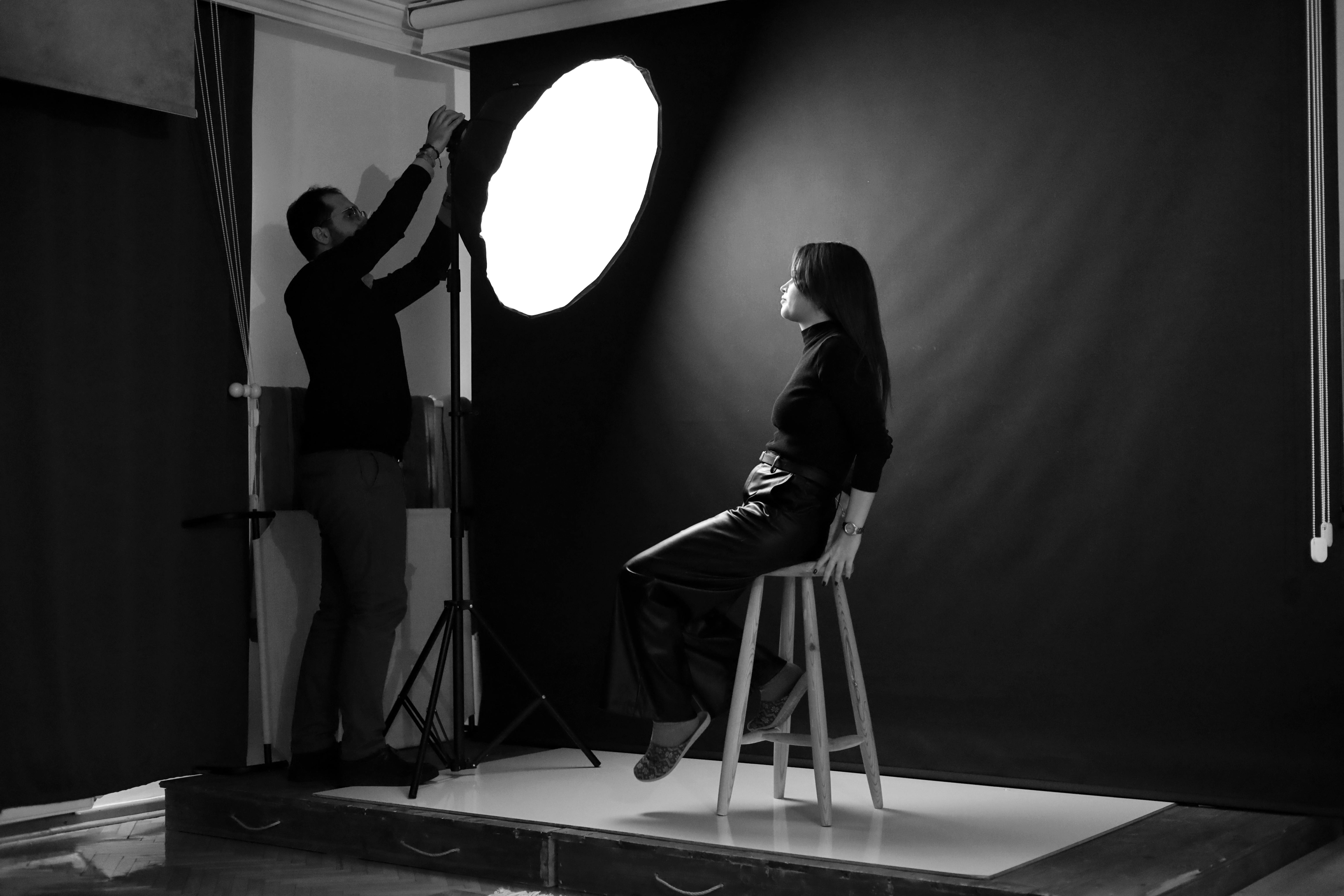 Free A photographer adjusts studio lighting for a female model seated on a stool. Stock Photo