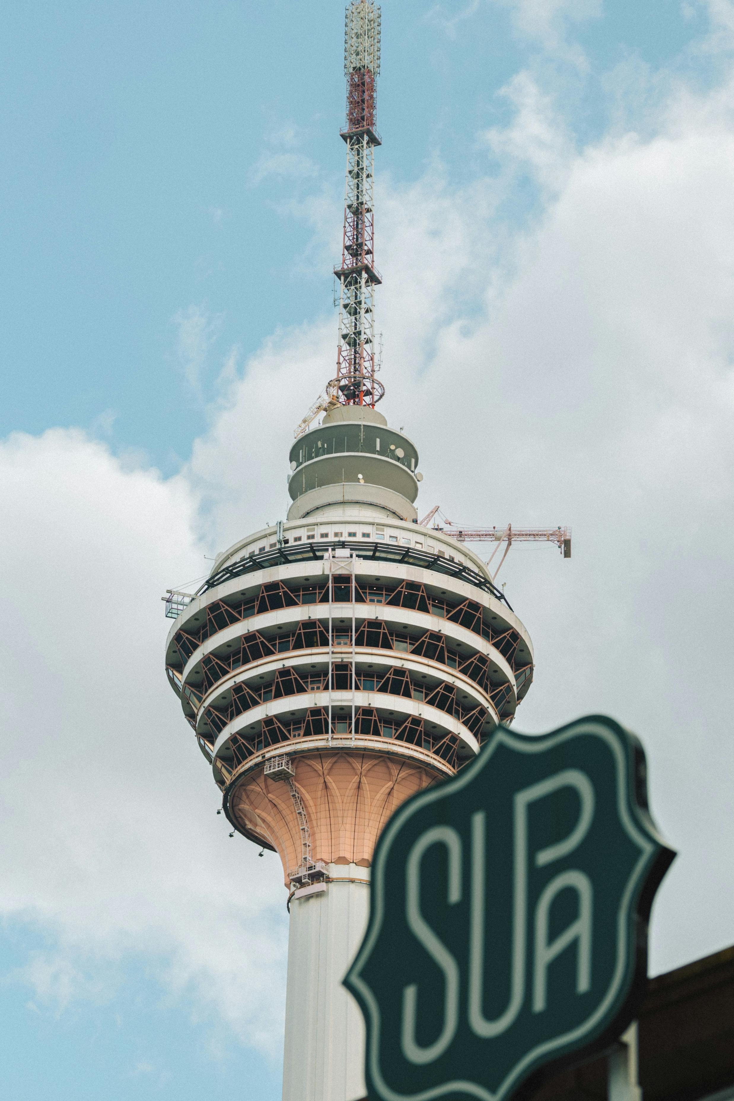 Gratis La icónica Torre de Kuala Lumpur se eleva contra un cielo azul nublado con un cartel de SURA en primer plano. Foto de stock