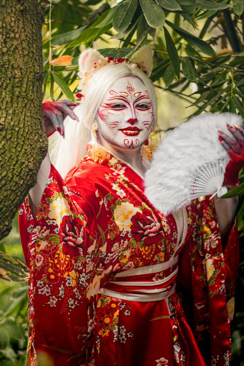 Free Woman in vibrant kimono with traditional makeup holding a fan in a lush park. Stock Photo
