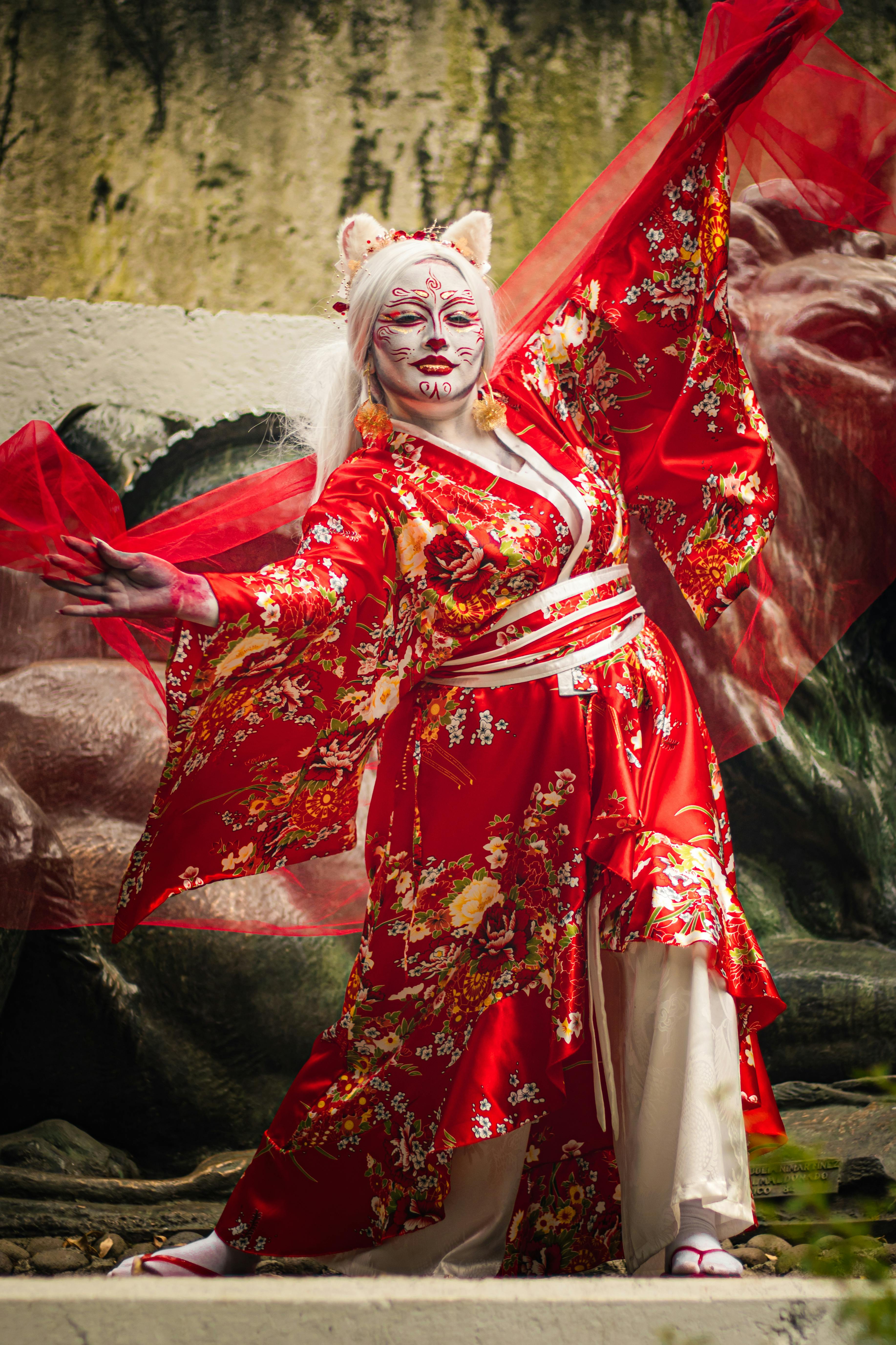 Ilmainen Punaiseen kimonoon pukeutunut esiintyjä taiteellisella meikillä luonnonkauniissa ympäristössä. Kuvapankkikuva