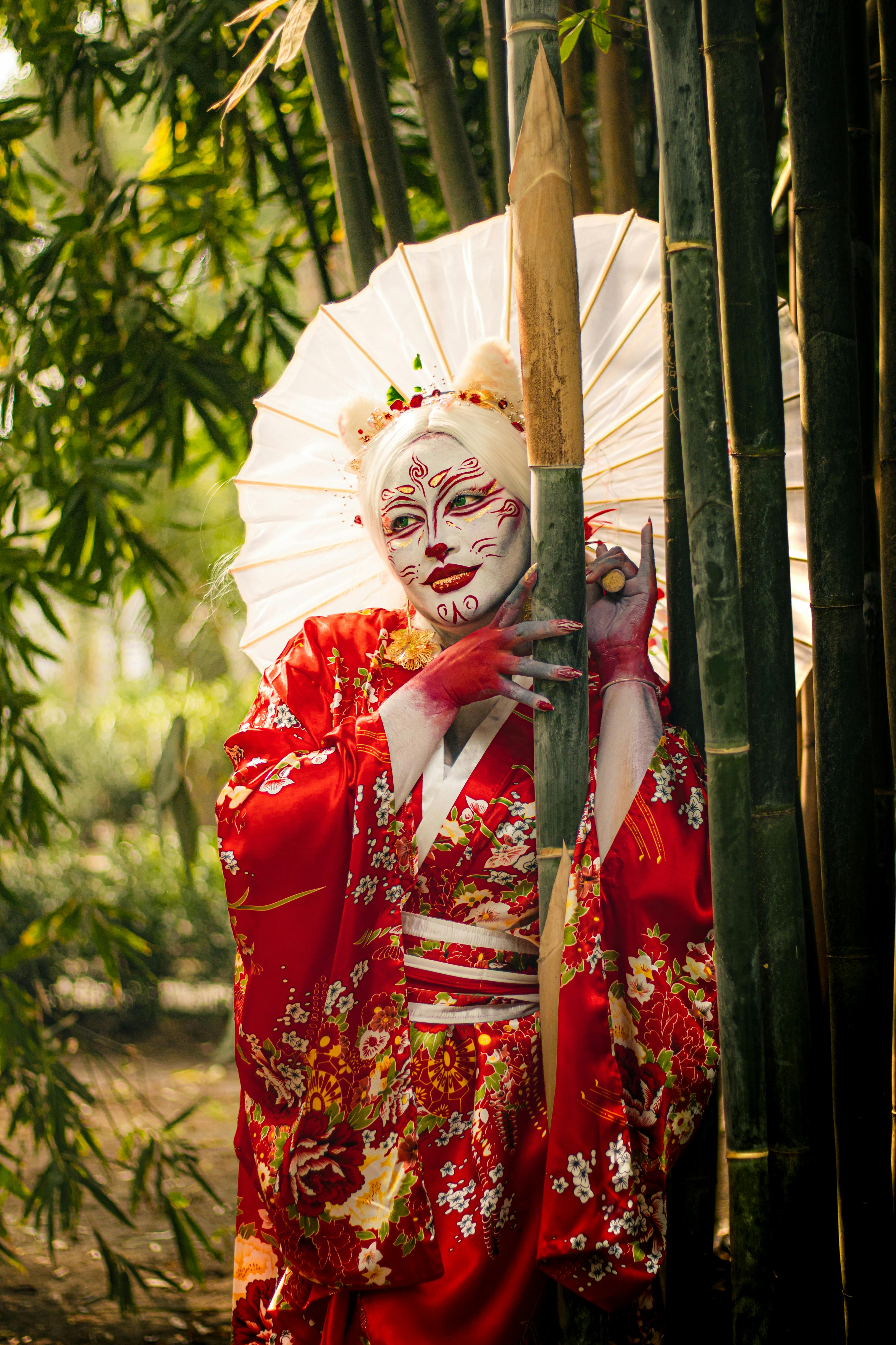 Ücretsiz Bambu korusunda, sanatsal makyajlı ve kırmızı kimono giymiş bir kişinin elinde şemsiye tutması, Japon kültür temalarını çağrıştırıyor. Stok Fotoğraflar