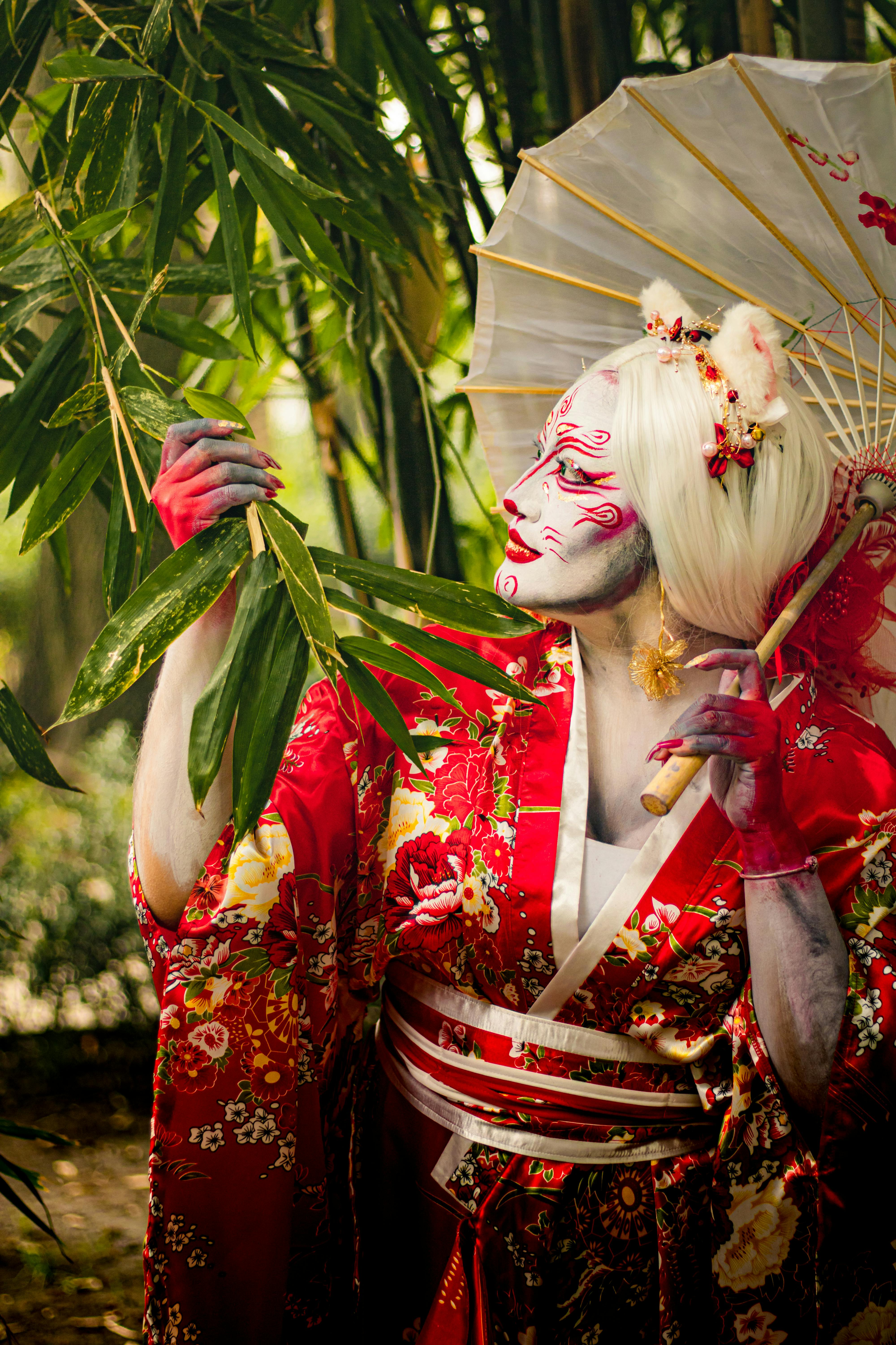ฟรี เครื่องแต่งกายเชิงศิลปะที่ได้รับแรงบันดาลใจจากเกอิชา พร้อมการแต่งหน้าจัดจ้าน และกิโมโนสีแดงแบบดั้งเดิม ในบรรยากาศสวนเขียวชอุ่ม คลังภาพถ่าย